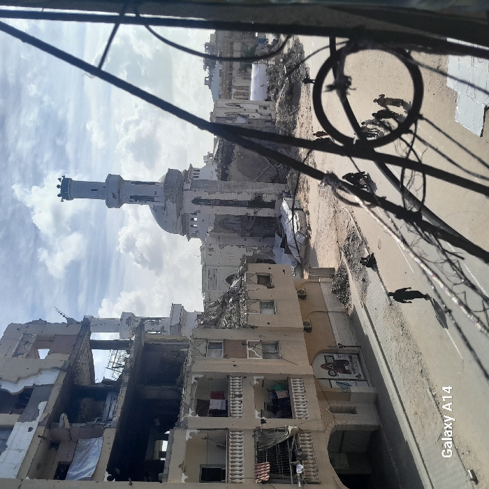 The image depicts a devastated urban area in Khan Yunis, Palestinian Territory, viewed from an elevated position, partially framed by dark metalwork. In the background, a large mosque stands severely damaged, with significant portions of its walls collapsed, revealing interior structures. Its tall minaret and central dome remain largely intact despite visible damage. To the left of the mosque, a multi-story building, likely residential or commercial, exhibits extensive structural damage, including collapsed floors, exposed rebar, and shattered facades; laundry is visible hanging on balconies of a lower, less severely damaged section.
The foreground features a paved street running horizontally, with a lone individual walking away from the viewer on its left side. Further down the street, several individuals are visible sitting or standing. The area between the paved street and the mosque is a wide expanse of disturbed earth, rubble, and sand, where a group of approximately 5-7 individuals walks towards the right. Scattered debris and remnants of structures are visible throughout the scene. The sky is partially cloudy. The bottom left corner of the image contains a "Galaxy A14" watermark.