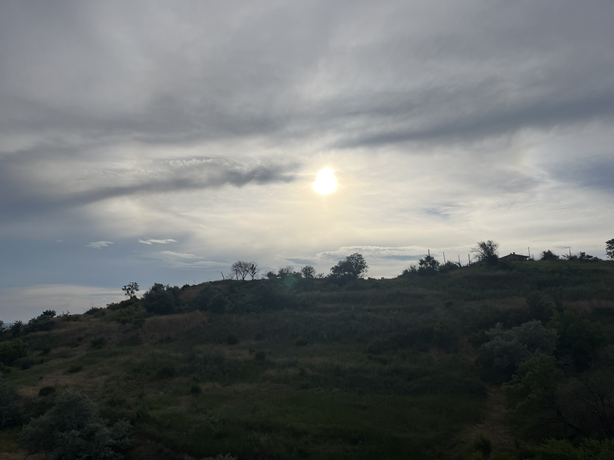 This is an outdoor, wide-angle photograph of a gently sloped, grassy hill with scattered trees and shrubs. The sky is overcast with dramatic clouds, and the sun is visible as a bright, glowing orb peeking through a gap in the clouds. The vegetation on the hill is a mix of dry grasses and greener bushes, with some bare trees visible on the crest. A small building with a red roof is perched on the top of the hill to the right of center. The scene appears to be a natural landscape, possibly on the outskirts of a city, with power lines visible on the hilltop. The lighting suggests it is either sunrise or sunset. The location is identified as the Asparuhovo district in Varna, Bulgaria, specifically in the area known as "Goliata" or "The Giant," a popular recreational spot with hiking trails and panoramic views.