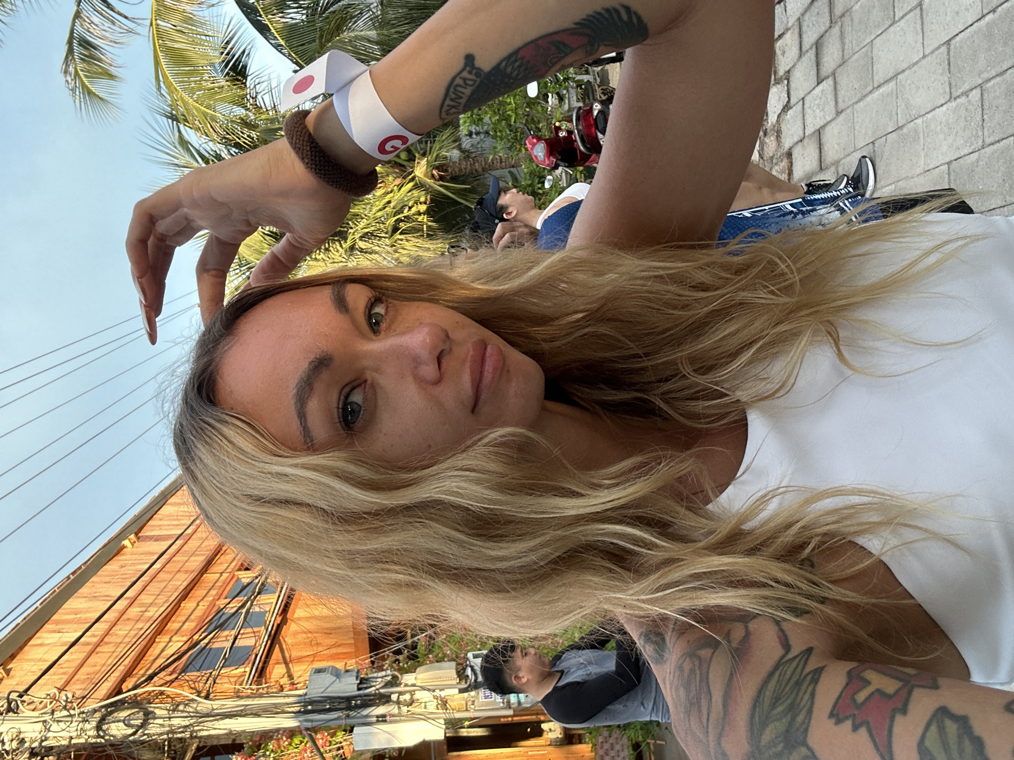 In Mũi Né, Vietnam, a blonde woman with tattoos on her arm and shoulder is taking a selfie. She is wearing a white tank top and has bracelets on her wrist, one of which is white with a red circle. Her right hand is raised to her head. Behind her, a man in a grey and black shirt stands near a building with an orange facade and visible electrical wires. Further in the background, palm trees, a red scooter, and other people are visible against a clear blue sky. The ground is paved with light-colored rectangular stones.