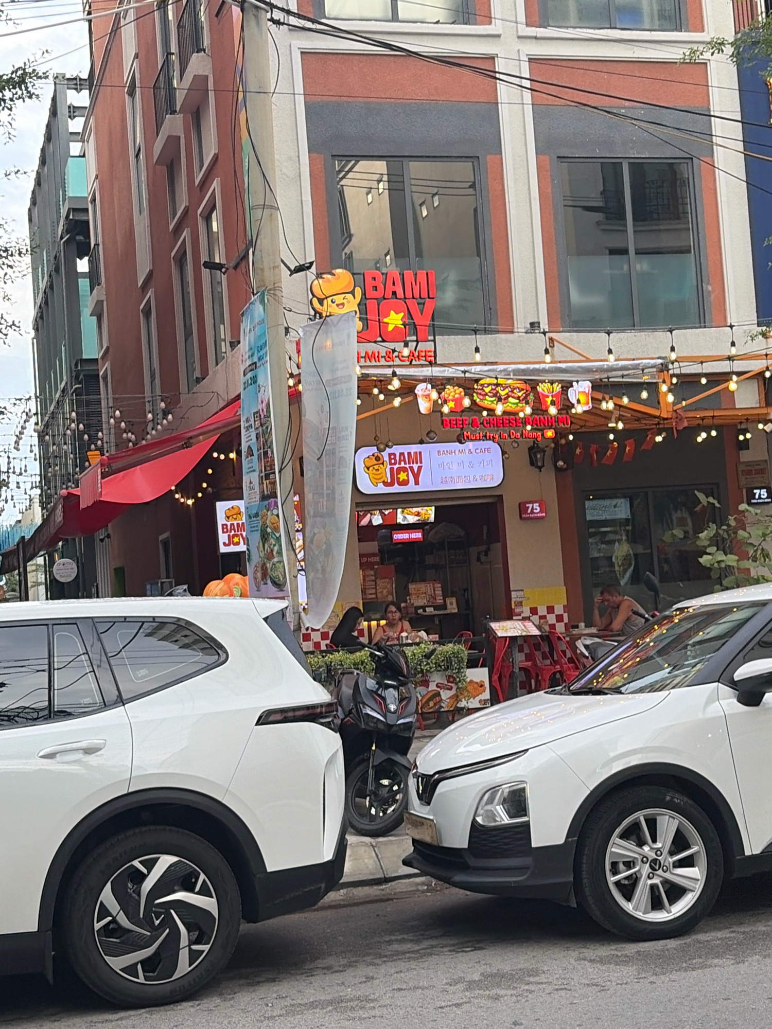 The image captures a street scene in Da Nang, Vietnam, featuring the exterior of a restaurant called "Bami Joy." The restaurant has a bright orange and red facade with a red awning. The name "Bami Joy" is prominently displayed in large, stylized letters. Above the entrance, strings of small lights are strung across, illuminating the area. Various food items, including burgers and drinks, are depicted in illuminated signage above the entrance.
In the foreground, the rear and side of a white SUV are visible, parked on the street. To the left of the SUV, a black motorcycle is parked. A woman is seated at a table outside the restaurant, facing away from the camera. Further to the right, another person is seated at a table, leaning forward with their elbows on the table.
The building itself appears to be multi-story, with windows and balconies visible on the upper floors. Electrical wires and poles are present along the street, typical of urban environments. Small Vietnamese flags are also seen hanging from the building, suggesting a nationalistic or celebratory theme. The overall impression is a bustling street view with a focus on a local eatery.