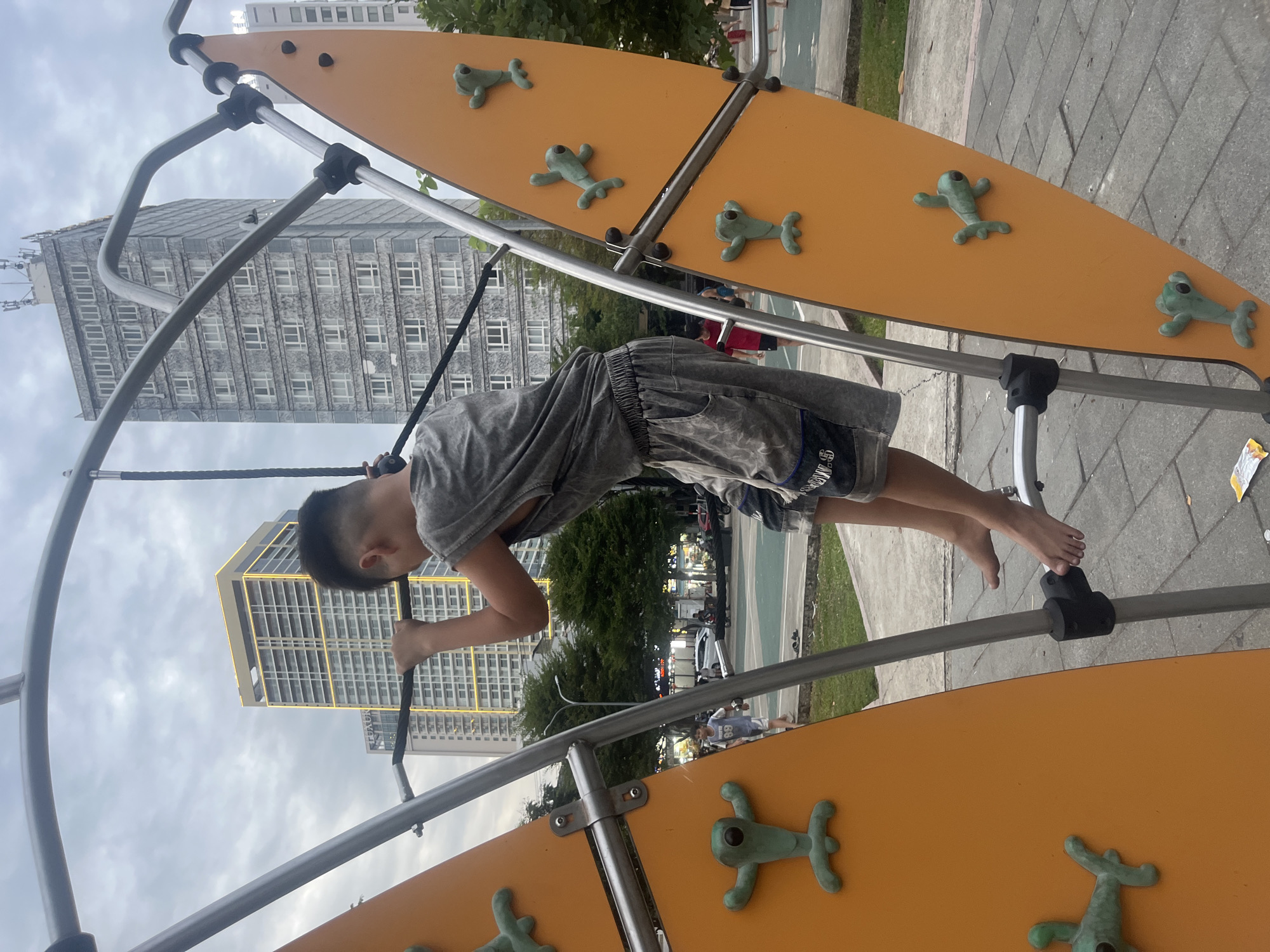 A young boy, appearing to be of East Asian descent, is actively climbing on an outdoor playground structure in a city park. He is wearing a grey sleeveless t-shirt and grey shorts. The playground structure consists of two orange panels with green, frog-shaped climbing holds, and a metal frame with ropes and bars. The boy is positioned in the center of the frame, his back to the camera, as he reaches for a horizontal bar above him. His left hand is firmly gripping the bar, and his right foot is placed on a metal rung.
In the background, several modern high-rise buildings are visible, characteristic of a city skyline. One building, with a yellow-lit top, stands out. The sky is overcast with clouds. The park area appears to be paved with brick-like stones and has green grassy areas. Other people, including children, can be seen in the background, suggesting a public recreational space.
The overall scene is indicative of a playground in Đà Nẵng, Vietnam, given the architectural style of the buildings and the presence of lush greenery. The time of day appears to be late afternoon or early evening, based on the lighting.