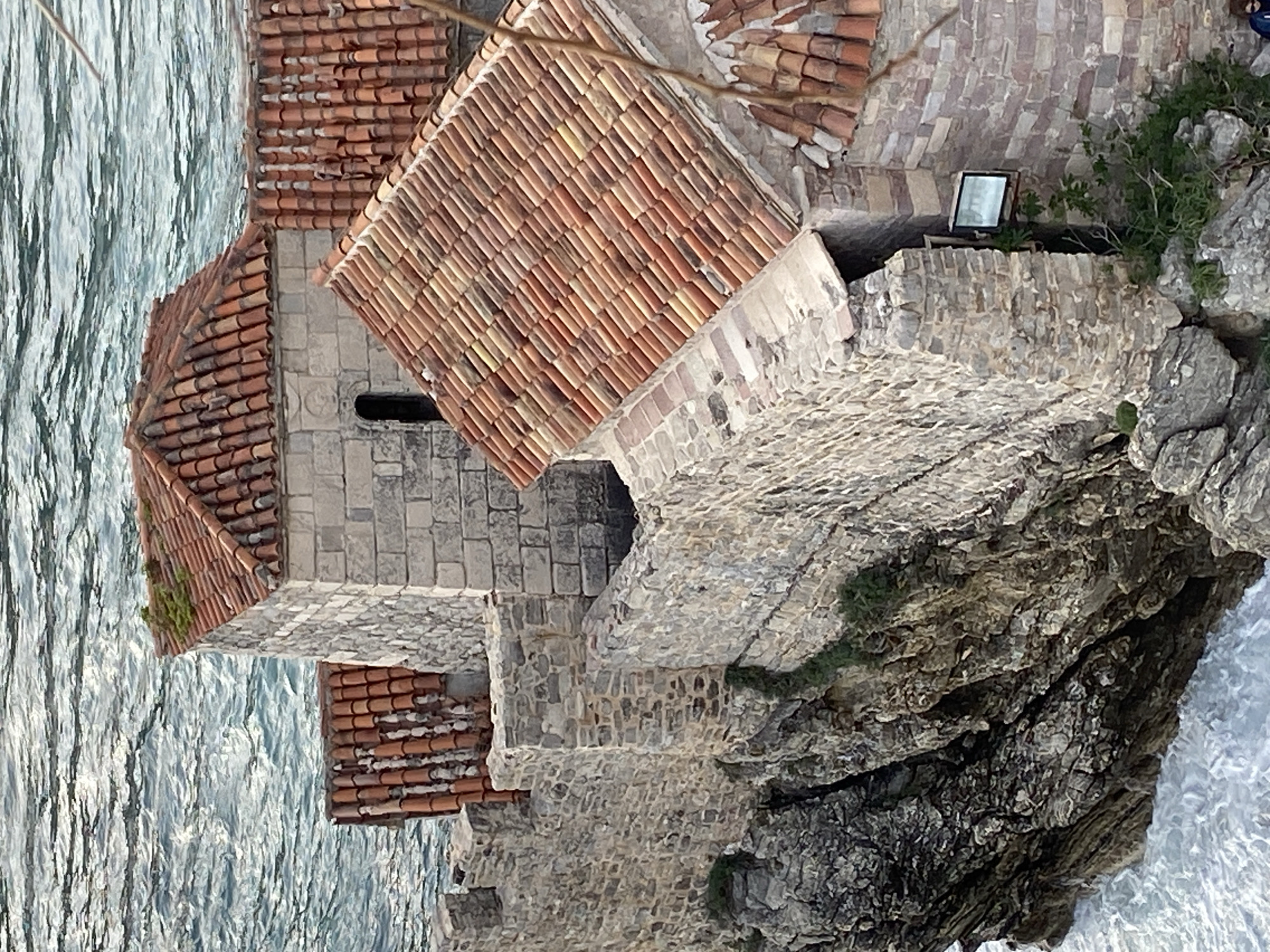 The image captures an aerial view of buildings with red-tiled roofs situated on a rocky coast, likely in or near Budva, Montenegro, based on the architectural style and coastal setting. The architecture consists of stone buildings with roofs made of uniform red clay tiles, typical of the Mediterranean region. Adjacent to the buildings is a rugged, rocky coastline. The sea extends towards the left of the frame, with the water surface showing subtle waves and reflections. A small, flat, possibly reflective object, perhaps a solar panel or mirror, sits on a flat stone surface. The perspective suggests the photograph was taken from an elevated position, possibly a higher building or a drone.