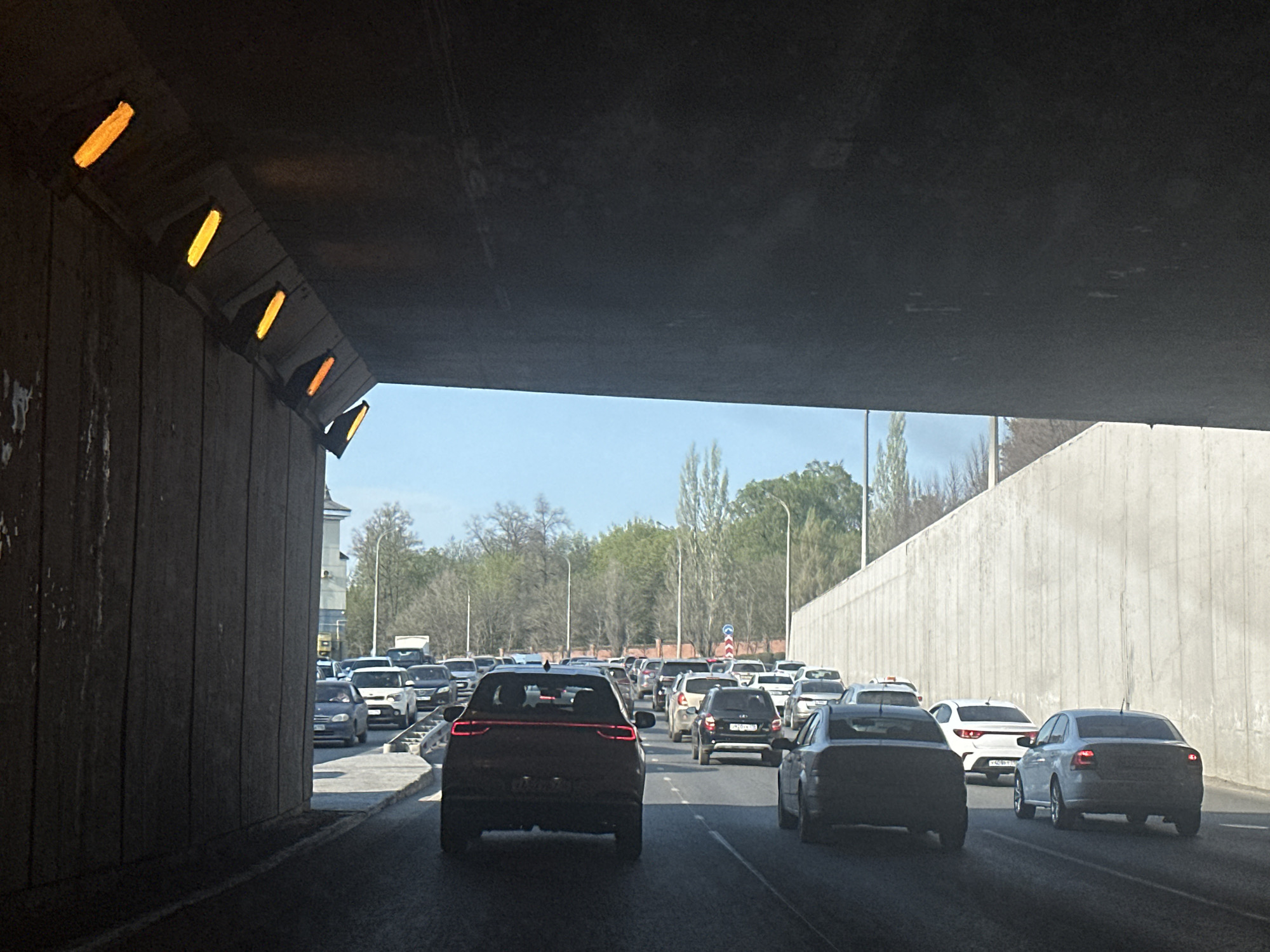 The image depicts a traffic jam inside a tunnel in Kazan, Russia. The tunnel walls are concrete, with orange rectangular lights affixed to the left side. Several cars are visible, mostly sedans and SUVs, driving slowly or stopped. Outside the tunnel opening, trees and buildings are visible under a bright, partly cloudy sky. The perspective is from inside a car, looking ahead through the tunnel.