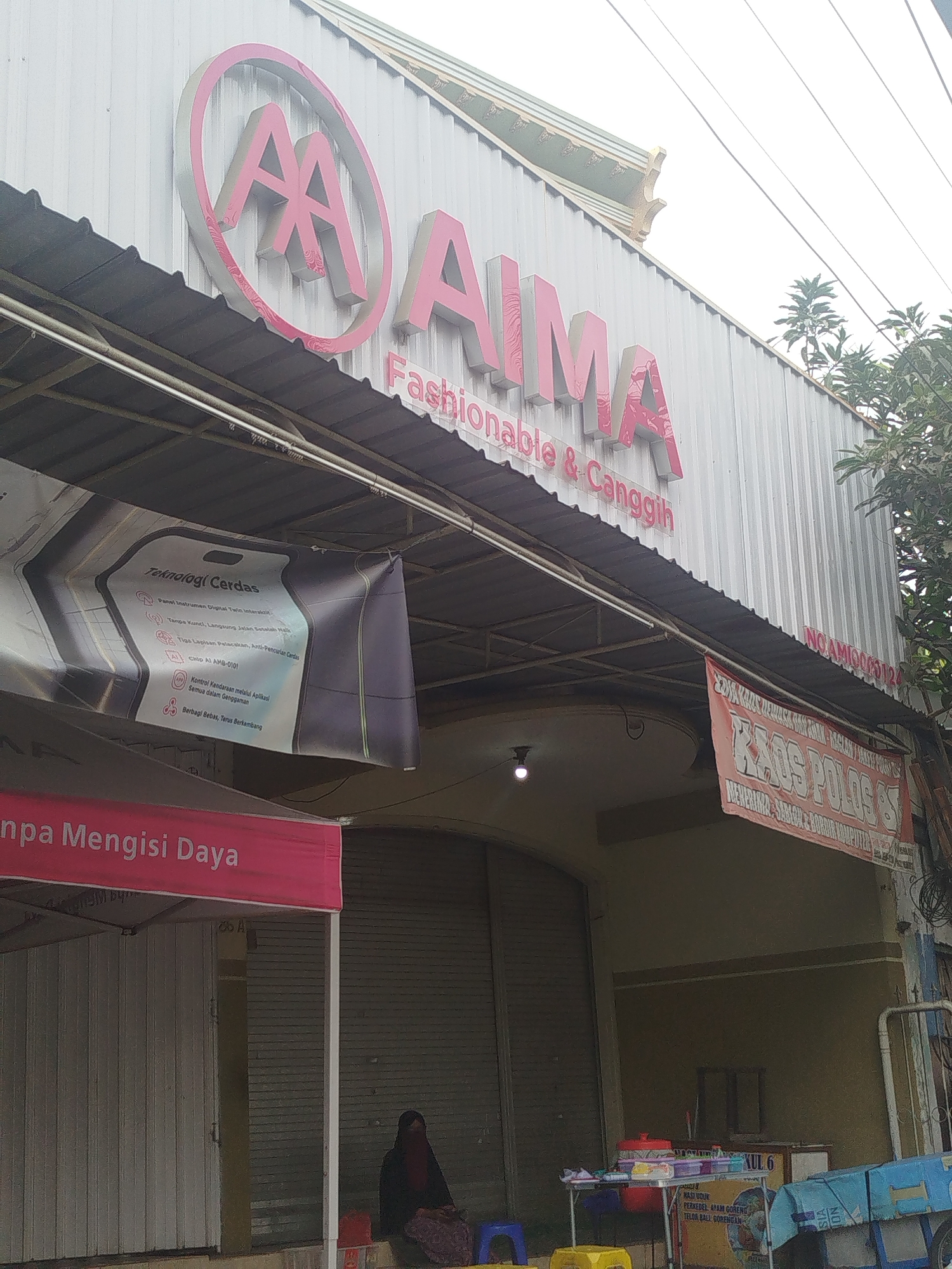 The image shows the facade of a commercial building in Yosowilangun, Indonesia. A large sign with the brand name "AIMA Fashionable & Canggih" is prominently displayed on the upper portion of the building. Below this, a banner advertises "Teknologi Cerdas" (Smart Technology) with bullet points detailing features. Further down, another banner displays "KAOS POLOS 65" (Plain T-shirts 65) and mentions "MERKIRKA BARGA MERKIRKA". A person wearing a dark headscarf and dark clothing is seated on the ground to the right of the image, near a closed rolling door. Various street furniture, including tables and chairs, are visible in front of the building, along with what appears to be a food cart. The overall scene suggests a retail or service establishment in a street-level commercial area.