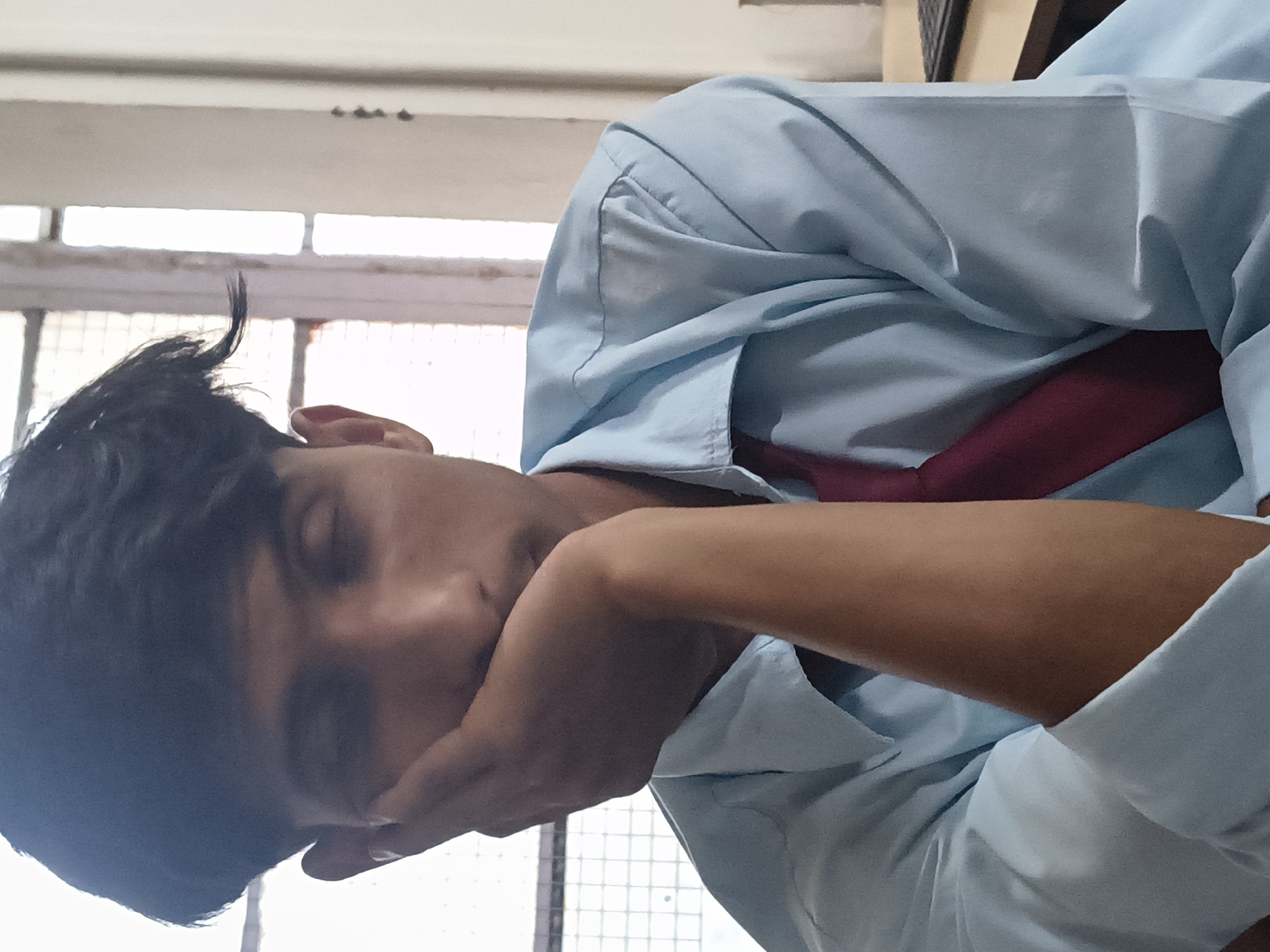A young man with dark hair rests his head on his right hand, his eyes closed, in what appears to be an indoor setting. He is wearing a light blue collared shirt and a dark red tie. The background suggests a room with a window that has a grid-like structure and vertical bars. To the right, a desk with a black keyboard is partially visible. The lighting is soft, coming from the front and the window, casting subtle shadows. The overall atmosphere conveys a sense of stillness and introspection.