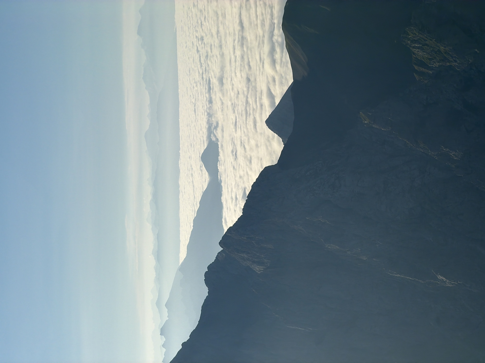 The image depicts a majestic mountain landscape viewed from a high vantage point. In the foreground, dark, rugged mountain slopes dominate the lower portion of the frame, showing deep shadows and textured rock faces. Above these, a vast expanse of fluffy white clouds stretches across the middle ground, creating a sea of mist that obscures the valleys below. In the distance, layer upon layer of fainter, blue-tinted mountain ranges recede into the horizon, softened by atmospheric haze. The sky above is a clear, pale blue, transitioning to a lighter hue near the horizon, suggesting either early morning or late afternoon light. The scene is devoid of any human presence or man-made structures, presenting a purely natural and serene environment.
While the image is a beautiful depiction of a mountain range, there is no specific visual information within the photograph that allows for identification of the location as Garmisch-Partenkirchen, Germany.