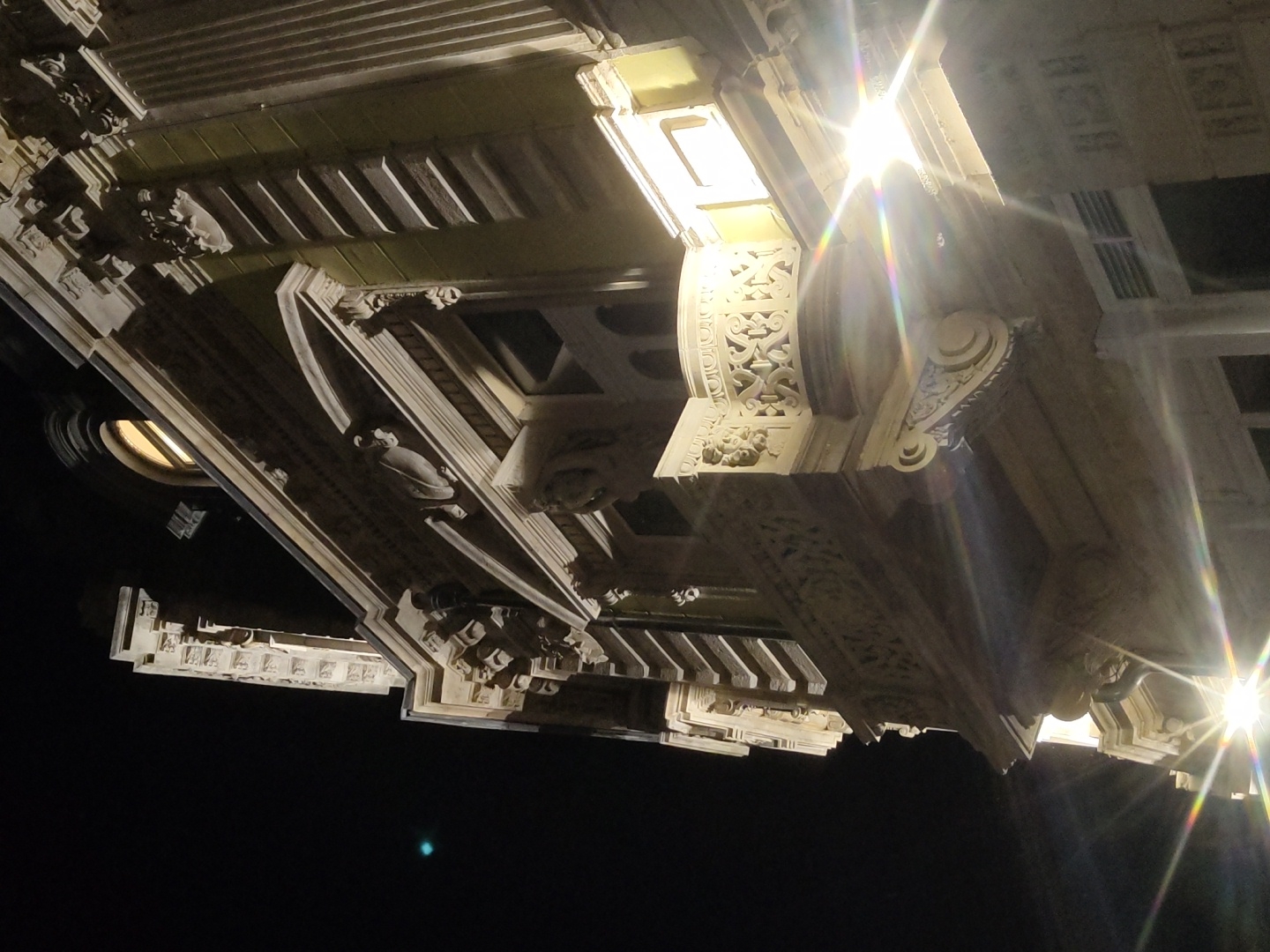 The image shows a low-angle night view of the facade of a building, likely a historical structure in Ruse, Bulgaria, characterized by ornate architectural details. The building features decorative carvings, cornices, and a balcony with intricate latticework. Artificial lighting illuminates the facade, creating bright highlights and lens flares. The sky is dark, indicating nighttime. The architectural style suggests a building from the late 19th or early 20th century, common in Ruse. No people are visible in the frame. The scene is static and focused on the building's exterior.
