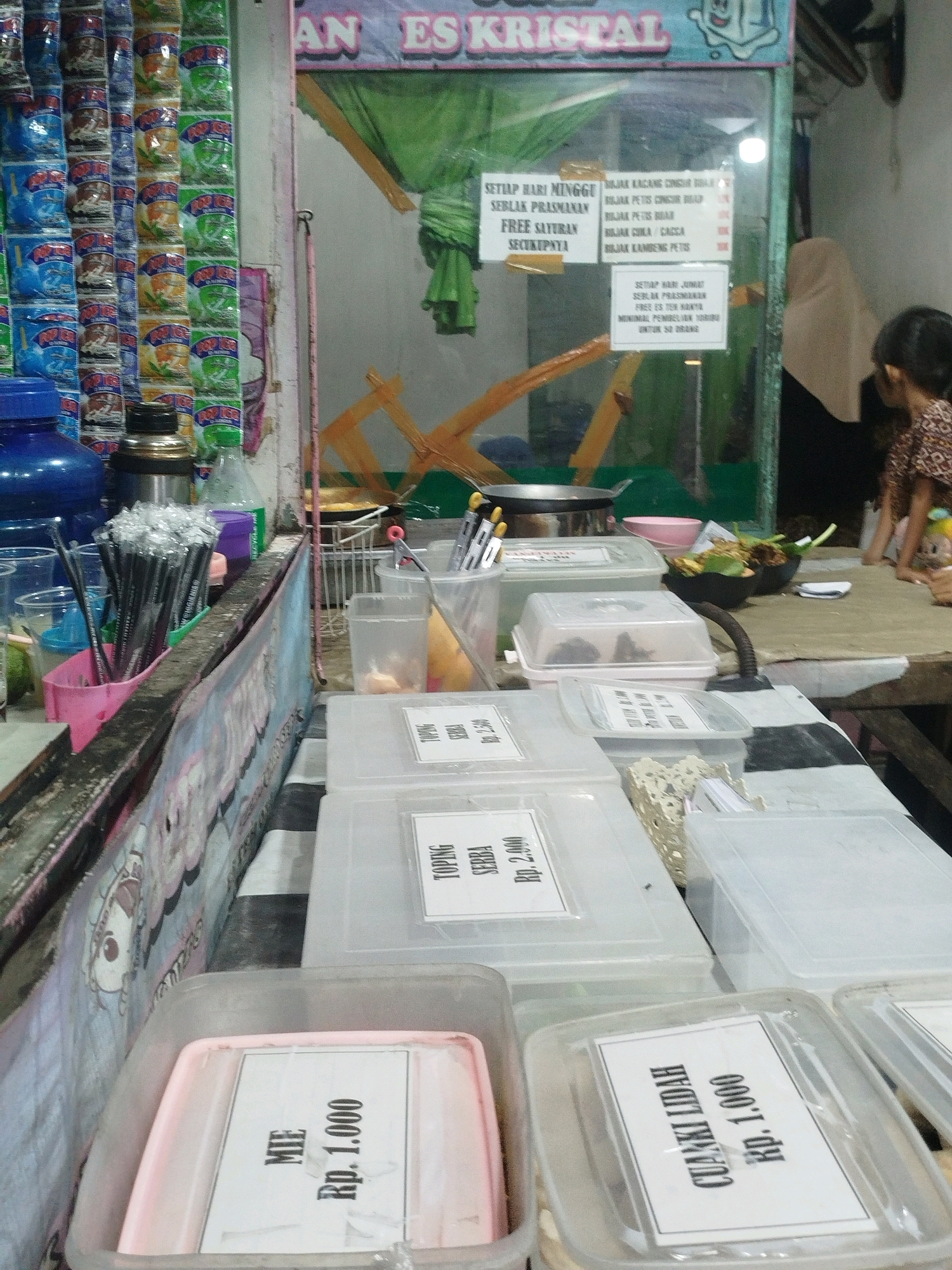 The image shows a street food stall in Burneh, Indonesia, with various food items and drinks on display. The stall is set up with a glass display case in the background, showcasing drinks like Es Kristal, and an assortment of packaged snacks. In the foreground, there are several clear plastic containers holding different food items, each with a label indicating the name and price. Some of these labels read "MIE Rp. 1.000", "TOPING SERBA Rp. 2.000", and "CUAKKI LIDAH Rp. 1.000". There are also bundles of packaged snacks hanging on the left side of the stall, and cooking utensils are visible. A woman wearing a hijab and a child are partially visible in the background, suggesting they are customers or staff. The overall scene depicts a typical outdoor food vendor operating in Indonesia.