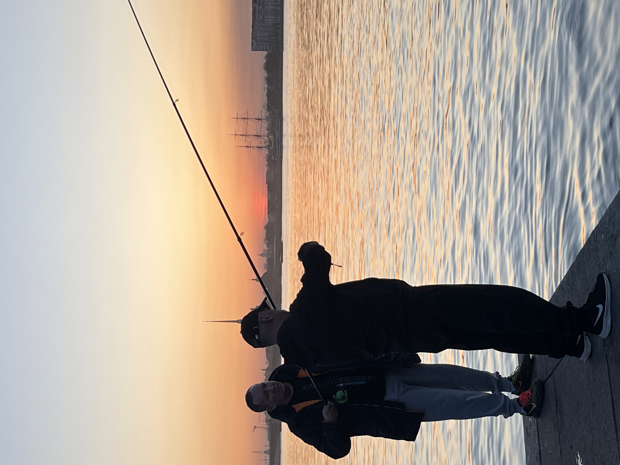 Two people stand on a wooden pier at sunset in Saint Petersburg, Russia, one holding a fishing rod. The cityscape is visible in the distance, including a sailing ship. The water reflects the warm colors of the sunset. The person closer to the camera is wearing a black jacket, gray sweatpants, and multi-colored sneakers. The other person is wearing a black cap, a black hoodie, black pants, and black sneakers with a white Nike logo. They both are looking toward the water.