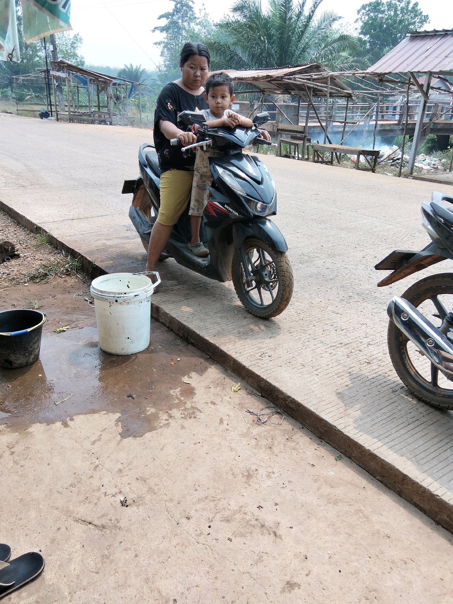 The image depicts a scene in Panca Desa, Indonesia, where a woman and a child are seated on a black Honda motorcycle. The woman, wearing a black t-shirt with a floral design and yellow shorts, is operating the motorcycle. The child, wearing a light-colored shirt and patterned trousers, is seated in front of her, holding onto the handlebars. They are positioned on a paved road, with a concrete barrier to their right. To the left of the motorcycle, a black bucket and a white bucket are visible on the ground, with some water spilled around them. In the background, there are wooden structures with corrugated metal roofs, suggesting a roadside market or resting area. Vegetation, including palm trees, can be seen further in the background. Another motorcycle is parked to the right of the main subject. The overall environment appears to be outdoors, in what seems to be a rural or semi-rural setting.