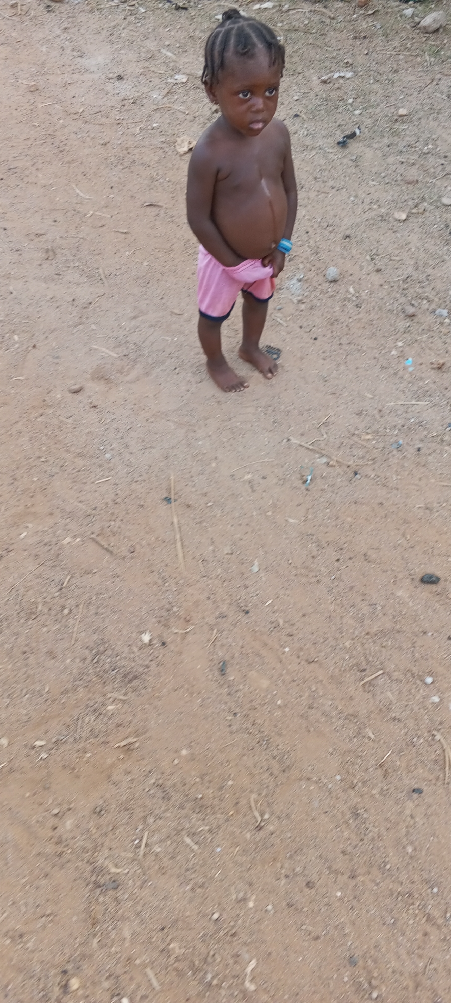A child stands on a dirt surface. The child has dark skin and is wearing pink shorts with blue trim. The child's hair is braided. The child is in the city of Keffi, Nigeria. The background is sandy with scattered debris.