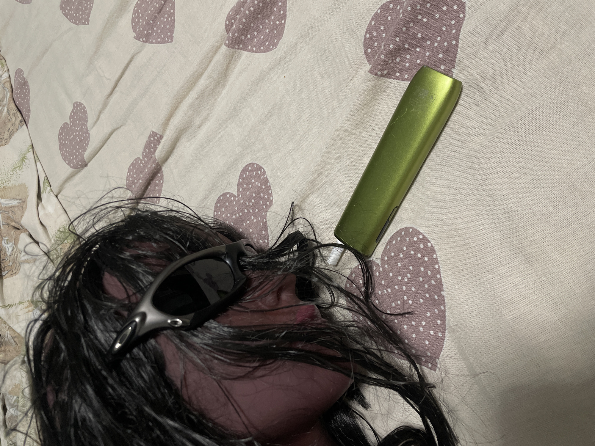 A close-up, overhead shot shows a detached head with black hair and dark sunglasses lying on a patterned comforter. A metallic green vape pen rests beside the head, near its mouth. The comforter has a repetitive pattern of pinkish hearts filled with white dots. The scene appears to be indoors, on a bed. There is no indication of location in the city of Unknown, Unknown.