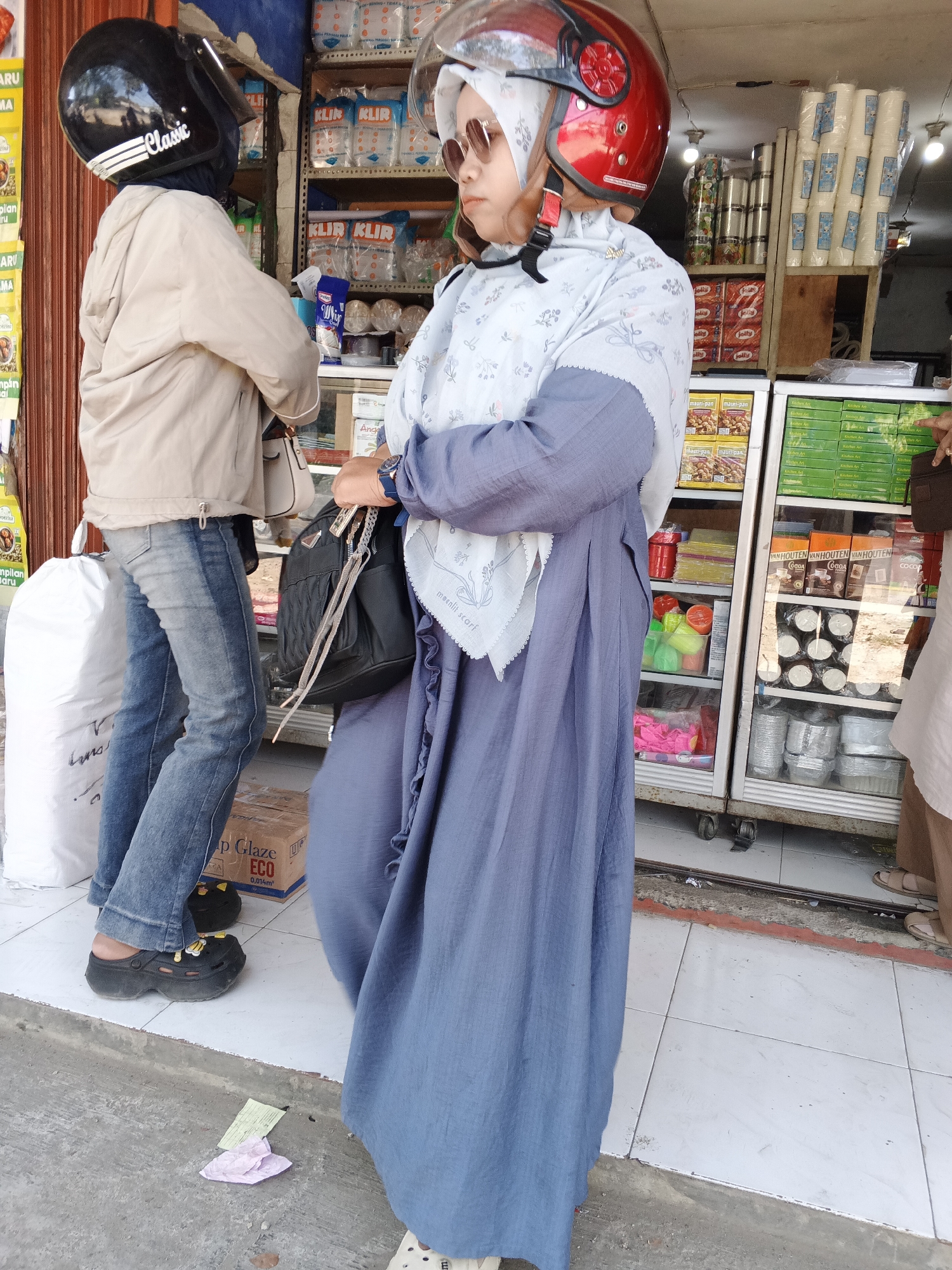 The image depicts two individuals standing outside a shop in Sukabumi, Indonesia. The person on the left is wearing a beige jacket, blue jeans, and black clogs. They are also wearing a black helmet with white stripes and the word "Classic" written on it. This person is partially obscured by the person on the right.
The person on the right is wearing a light blue hijab and a long, flowing blue dress. They have on sunglasses and a red helmet. They are carrying a black handbag with a chain. Both individuals appear to be facing the shop, which displays various products on shelves, including what seem to be food items and household goods. The shop has a metal shutter and is located on a tiled sidewalk. The ground in front of the shop shows some scattered papers and debris. The overall scene suggests a typical street-level commercial area in Indonesia.
