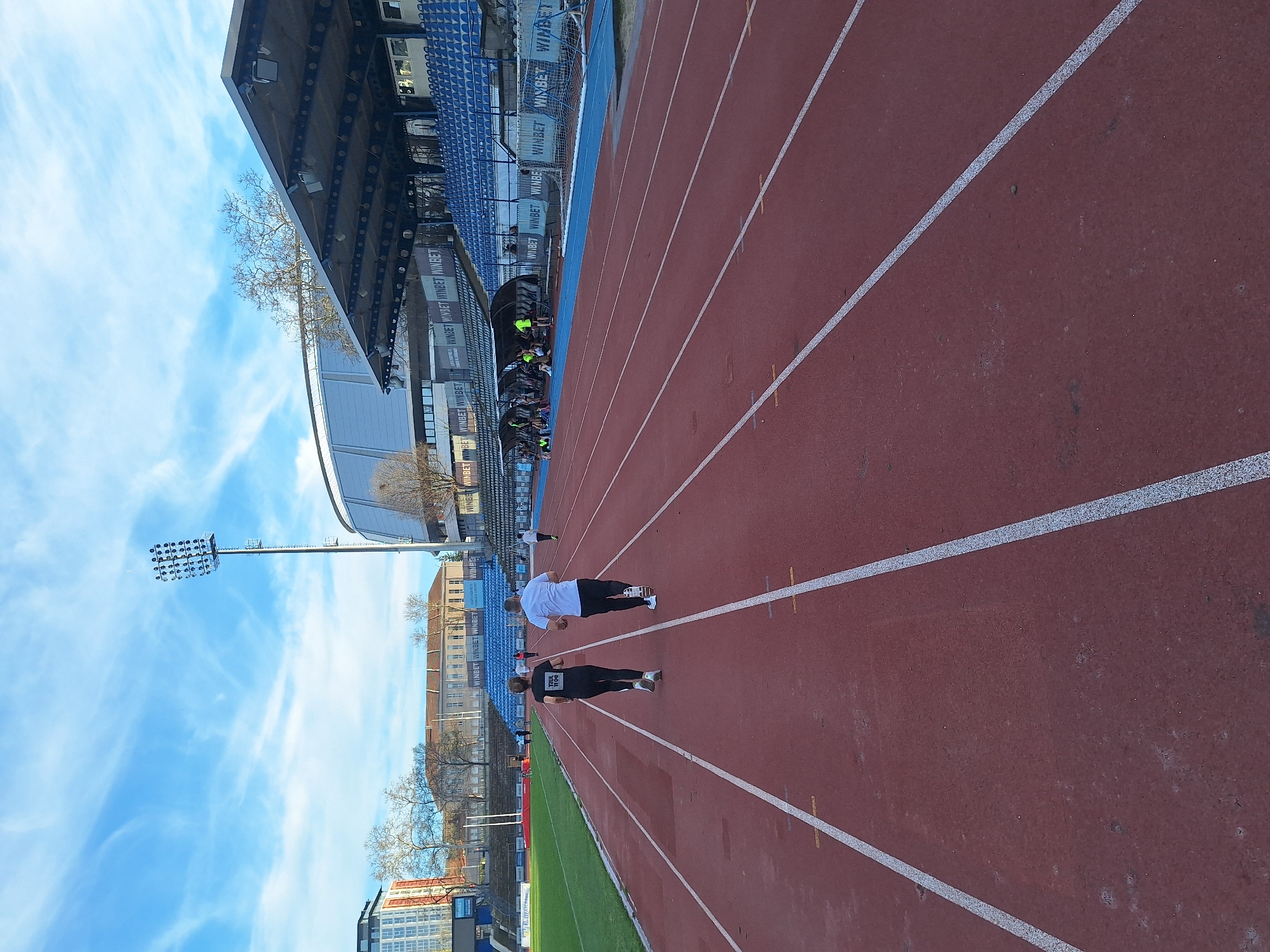 The image depicts an outdoor athletic track, possibly in the city of Unknown, Unknown. The track is red, with several white lane markers clearly visible. Two individuals are seen on the track: one wearing a white shirt and black shorts, and another wearing black athletic wear. They appear to be running or jogging. To the left of the track is a grassy area, followed by stadium seating under a structure. Banners advertising "WINBET" are prominent along the side of the stadium. A tall lighting structure is visible in the distance, reaching towards the blue sky with scattered clouds. Buildings and trees are also seen in the background.