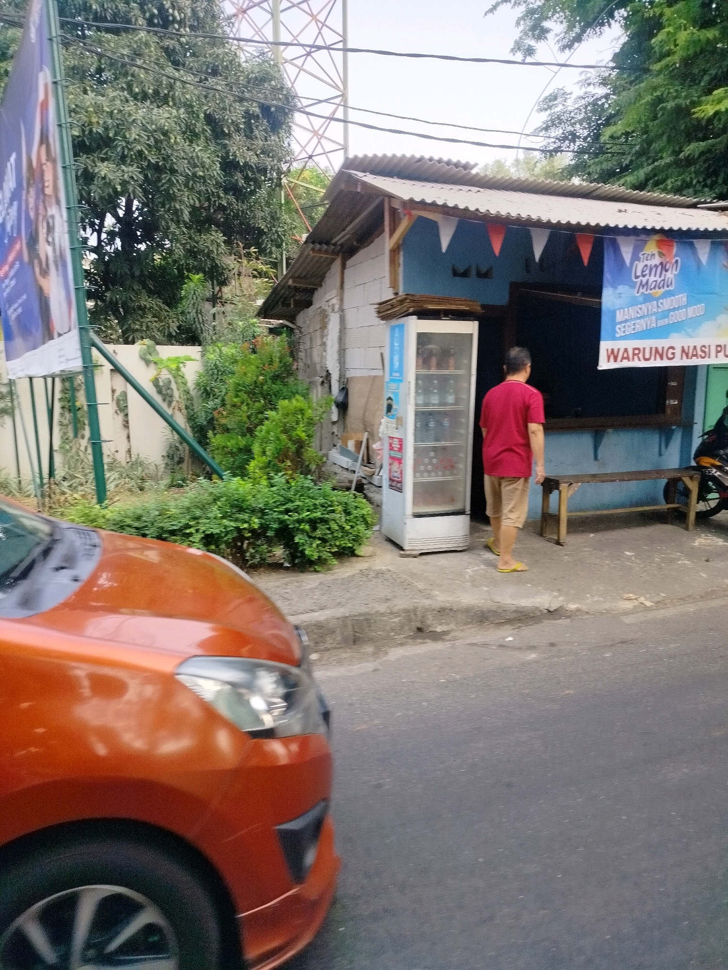 The image shows a street scene in East Jakarta, Indonesia, with a small roadside warung (stall). A man wearing a red t-shirt and tan shorts stands facing away from the camera, looking at a refrigerator filled with drinks. To the left of the warung, a large blue banner advertises "Teh Lemon Madu" and "WARUNG NASI PU". A wooden bench sits in front of the warung. An orange car is visible in the foreground, partially obscuring the street. Green foliage and trees are present in the background, along with a tall metal structure that appears to be a utility tower. The warung has a corrugated metal roof and a light blue wall. The ground is paved with concrete in front of the warung and transitions to asphalt on the street.