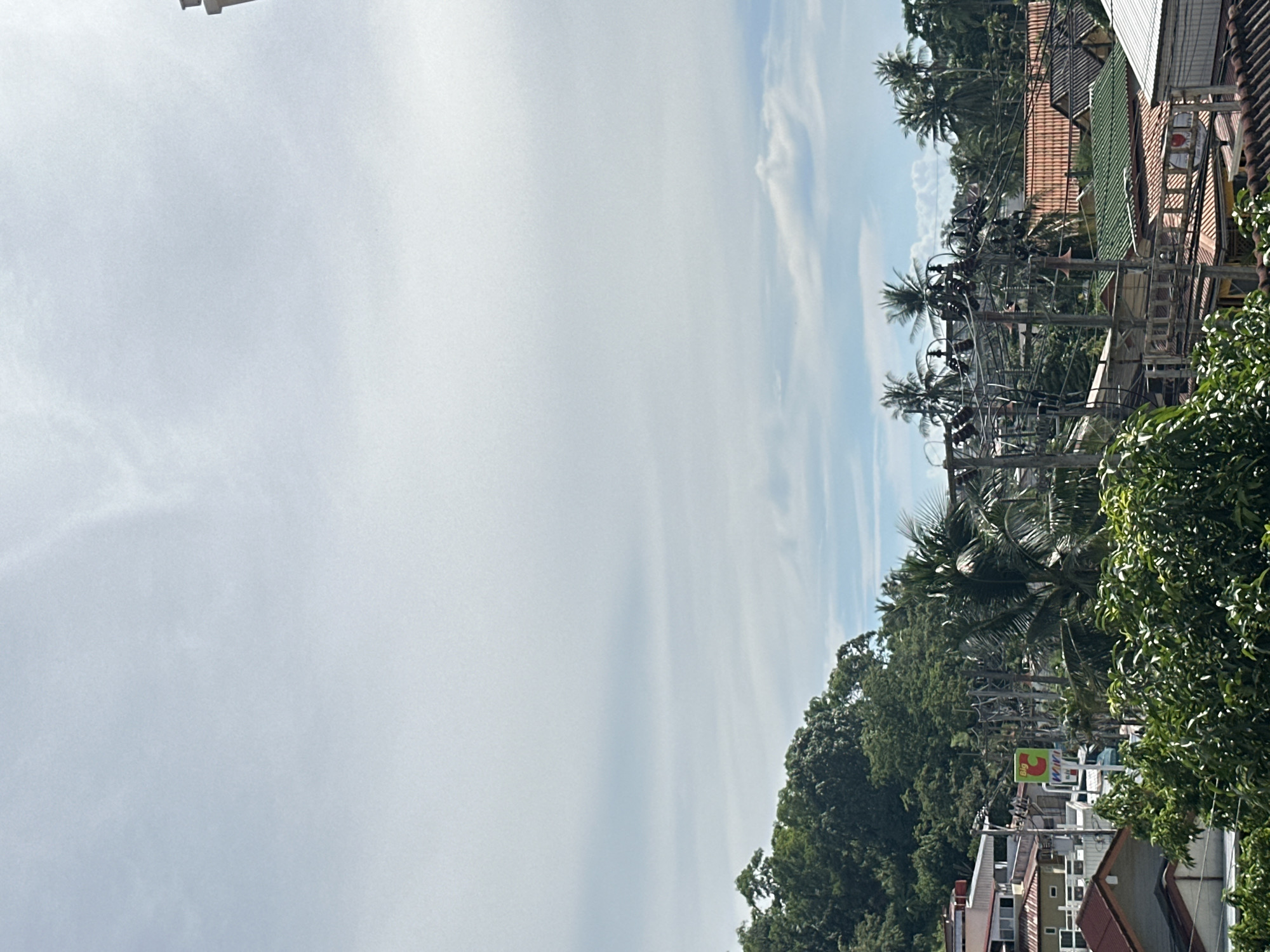 The media file is a vertical, eye-level shot of a street scene in Karon, Thailand. The sky is a pale blue with wispy white clouds, suggesting a clear day with some cloud cover. The right side of the frame is dominated by lush green trees and palm trees, indicative of a tropical environment. Interspersed among the greenery are buildings with terracotta-colored tiled roofs, typical of residential or commercial structures in the region.
A prominent feature is a dense network of utility poles and wires crisscrossing the frame, suggesting a busy urban or semi-urban area. These poles are laden with electrical wires, creating a complex, almost tangled appearance. In the distance, a few buildings are visible, some with white or light-colored facades.
A notable detail is a green sign with a red "3" and yellow "Big C" logo, which is a well-known supermarket chain in Thailand. This sign provides a clear location cue. The overall impression is of a sunny day in a vibrant, potentially bustling, Thai town, with a mix of natural tropical elements and man-made infrastructure. No people are visible in the frame, and the focus is on the streetscape and its surroundings.