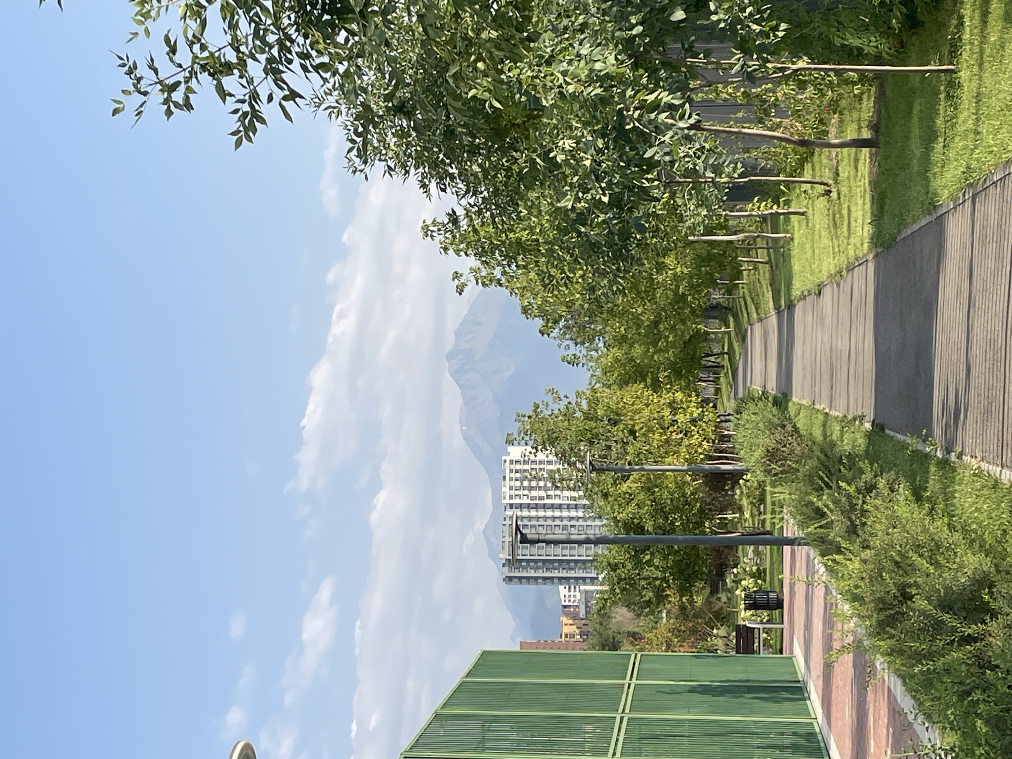 The image depicts a view down a paved pathway lined with young trees and manicured bushes, likely in a park or residential complex in Almaty, Kazakhstan. The pathway leads towards a backdrop of modern apartment buildings and, further in the distance, the majestic, snow-capped Zailiysky Alatau mountains. The sky is a clear blue with scattered white clouds. To the left of the pathway, a tall, green, slatted fence encloses an area, and beyond it, red brick paving and a bench are visible. Lampposts line the pathway, indicating it is designed for walking and possibly evening use. The overall impression is one of a well-maintained urban green space with a striking natural landscape as its distant feature.