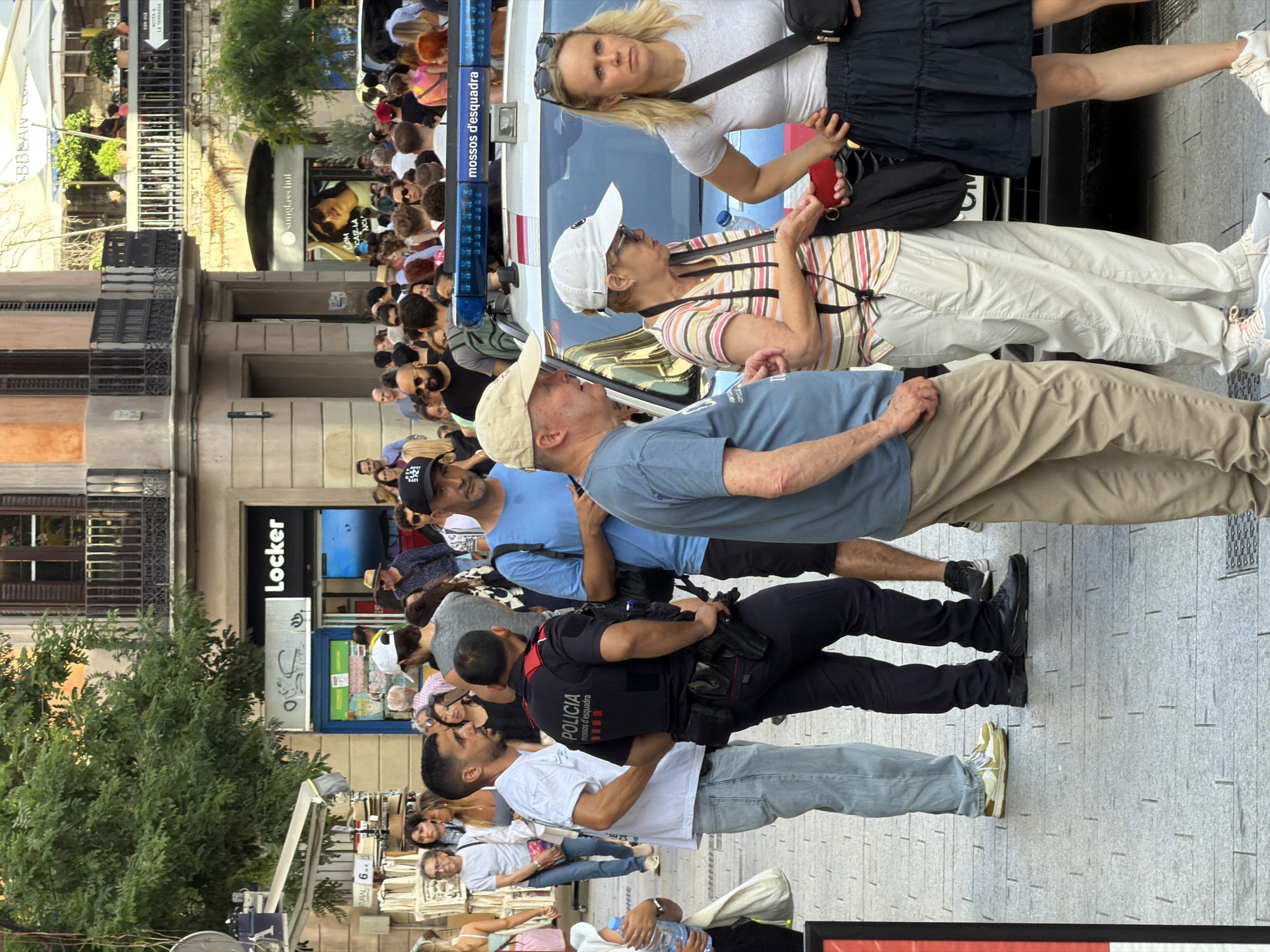 This image captures a street scene in Barcelona, Spain, likely near a shopping area, given the presence of storefronts. In the foreground, a group of people are gathered. A police officer in uniform, identified by the text "POLICIA MOSSOS D'ESQUADRA" on his back, is standing with his hands behind his back. He is wearing a dark uniform with a utility belt. To his left is a man in a white t-shirt and light-colored jeans. To the officer's right are several civilians: a man in a light blue t-shirt and khaki pants, an older woman in a striped shirt and light-colored pants, and a woman with blonde hair wearing a grey t-shirt and a dark skirt. A police vehicle with flashing blue lights is visible behind them, with "mossos d'esquadra" written on the light bar. In the background, there are more people walking and standing, and various building facades, including a "Locker" store. Trees and awnings suggest an urban outdoor environment. The overall atmosphere appears to be one of a busy public space, with a police presence.