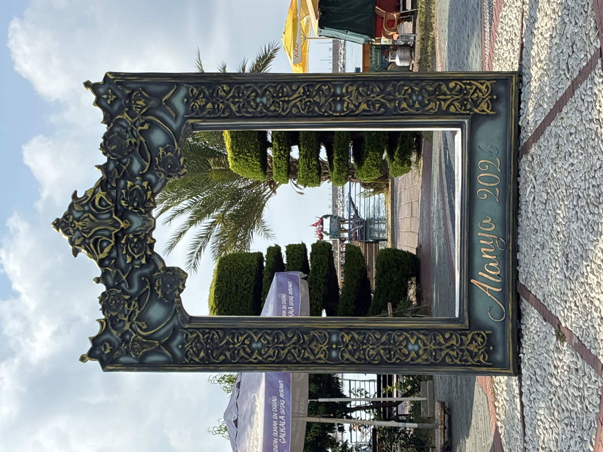 A large, ornate, dark green and gold picture frame stands on a pebbled walkway in Alanya, Turkey. The frame is decorated with intricate scrolling patterns and floral motifs. Through the frame, a view of the Alanya harbor and coastline is visible. The sky above is partly cloudy. The frame has the words "Alanya 2026" inscribed in gold lettering on its lower section. In the background, palm trees, manicured hedges, and umbrellas associated with outdoor seating areas can be seen. A boat is docked in the harbor. The overall scene suggests a public promenade or tourist area.