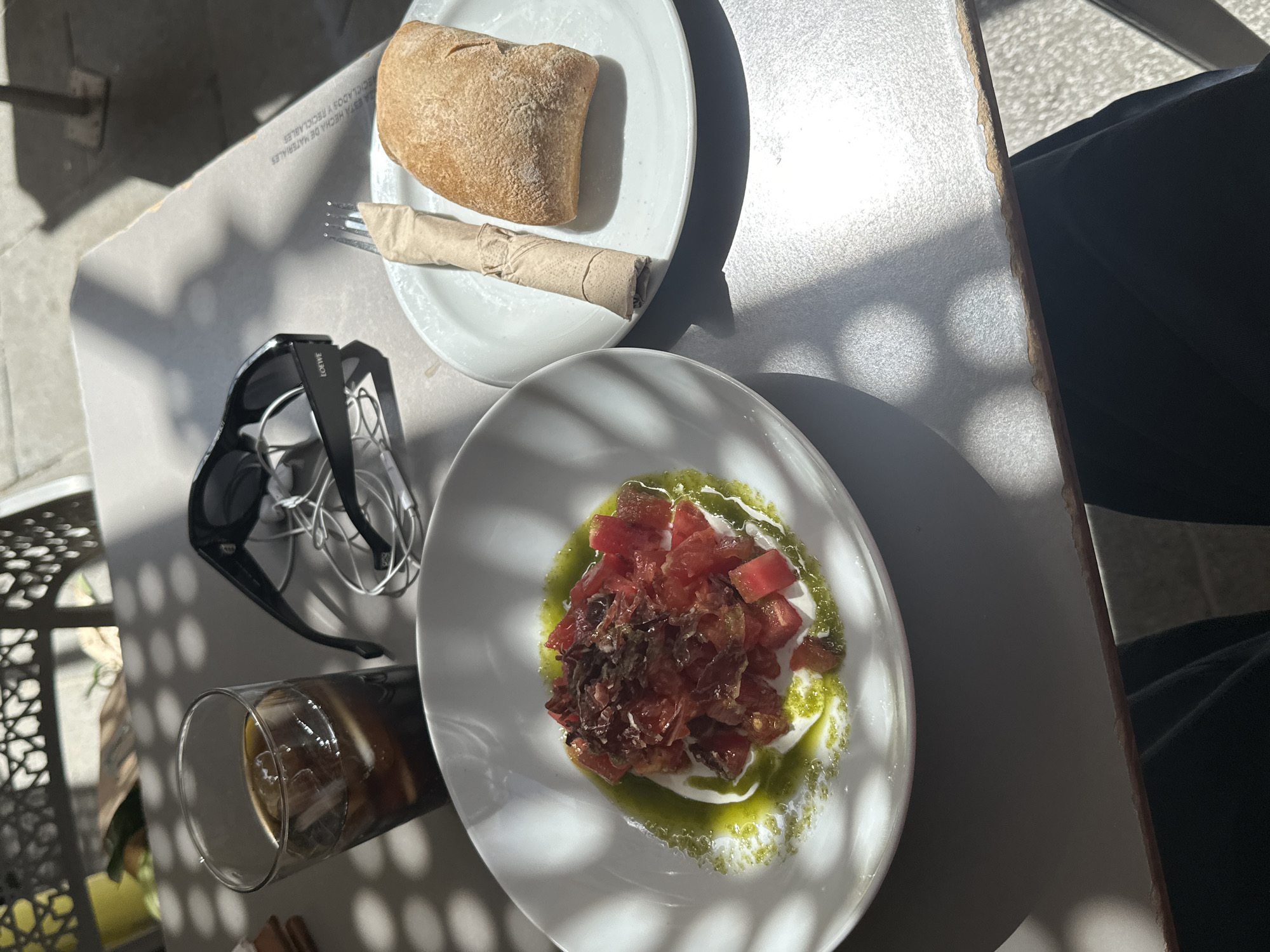 The image displays a bright, outdoor dining scene, likely in Madrid, under direct midday sunlight that casts strong, patterned shadows across the table. A light-colored table is set with a casual meal for one.
On the left, a white plate holds a rustic bread roll and a wrapped fork in a brown paper napkin. This plate rests on a recycled cardboard placemat, which bears Spanish text that translates to "IT IS MADE OF RECYCLED MATERIALS AND RECYCLABLE." Beside the plate, a pair of black sunglasses with "LOEWE" visible on the arm rests near tangled white earbuds. A tall glass contains a dark iced beverage, possibly coffee or cola, with ice cubes.
The main dish is presented on a white oval plate, featuring diced red tomatoes, a creamy white base that appears to be burrata or mozzarella, and a vibrant green oil or pesto drizzle. Topping the dish are shredded dark pieces, strongly resembling jamón serrano, a common Spanish cured ham.
The distinct shadows suggest an overhead trellis or sunshade. A patterned metal chair is partially visible on the left, and a person's leg in dark trousers is visible on the right, indicating someone is seated at the table enjoying the meal. The overall atmosphere is relaxed and summery, typical of an outdoor café setting.