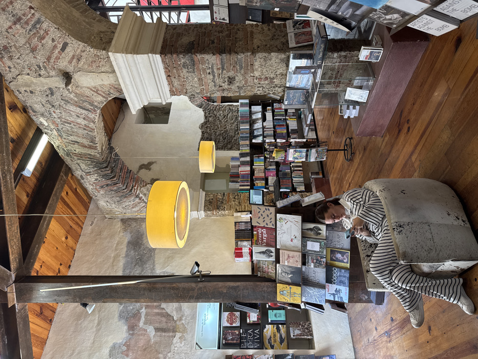 The image depicts the interior of Profética, Casa de la Lectura, a bookstore located in the historic center of Puebla, Puebla, Mexico. The environment features a rustic aesthetic with exposed wooden ceiling beams and planking, irregular plaster walls showing sections of exposed brick and stone, and polished wooden floorboards. Structural elements include large dark wooden columns and a prominent brick archway supporting the upper structure.
Illumination is provided by two large, circular yellow fabric pendant lamps hanging from the ceiling, augmented by fluorescent strip lighting and track lights mounted on the wooden beams.
A single female individual is seated in the foreground, slightly right of center. She is a young adult with light skin and dark hair, wearing a black and white striped long-sleeved shirt and matching wide-leg pants, with white sneakers. She sits in a low, rounded armchair upholstered in a textured, light-colored fabric, possibly cowhide. Her focus is on a smartphone held in both hands, which she is actively operating. Her legs are crossed at the ankles.
The bookstore is densely populated with books displayed on various shelving units, tiered racks, and in glass-topped display cases. Titles visible include "Piedra Viva," "Gerardo Vargas," and "Código," indicating Spanish-language publications. A security camera is mounted on a wooden column to the left. In the background, beyond the brick arch, a large window or opening with a distinctive red frame is visible, hinting at an exterior view or an adjoining section of the building.