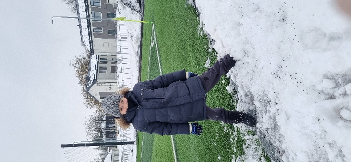 An individual child stands in an outdoor sports facility in Pavlovsk, Russia, under overcast winter conditions with partial snow cover. The child, estimated to be between 8 and 12 years old, is positioned centrally, facing slightly left of the camera, with a faint smile. The child wears a grey knitted beanie, a dark grey or navy quilted winter jacket with a fur-trimmed hood, dark trousers, blue and white striped gloves, and black boots. A dark neck gaiter or scarf covers the lower portion of the face.
The immediate foreground consists of a mound of freshly accumulated snow. The child's left foot is embedded in this snow, while the right foot rests on a green artificial turf football pitch. White lines delineate the pitch, which is partially covered by snow, especially along its perimeter. A yellow corner flag is visible on the right side of the pitch. To the far left, a dark net structure, possibly part of a goal, is partially visible. In the background, several low-rise buildings with grey facades and dark roofs are present, interspersed with bare deciduous trees and some still retaining brown leaves. A streetlamp pole is visible on the right side of the background. The sky is uniformly grey.