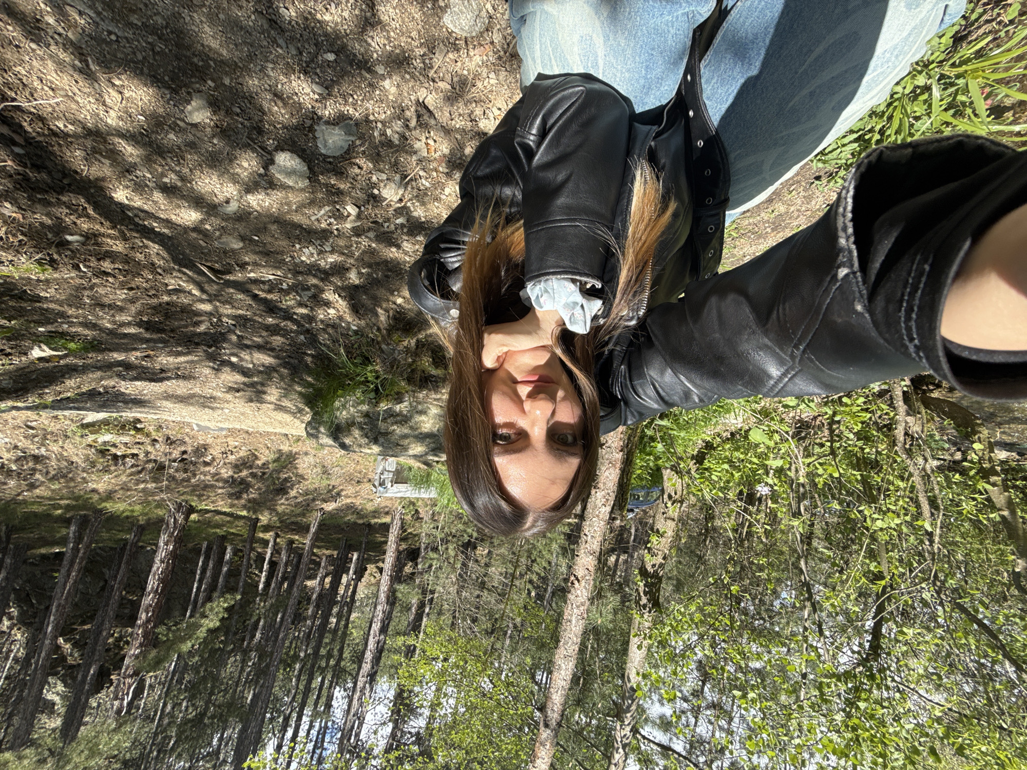 A young woman wearing blue jeans and a black leather jacket is positioned in the center of the frame. The image appears to have been taken at an unconventional angle, causing the trees in the background to appear as though they are growing "downwards." The background displays a forest with tall, thin trees. To the left, there is a dirt ground covered with leaf litter and small rocks. The location is Ovchartsi, Bulgaria.