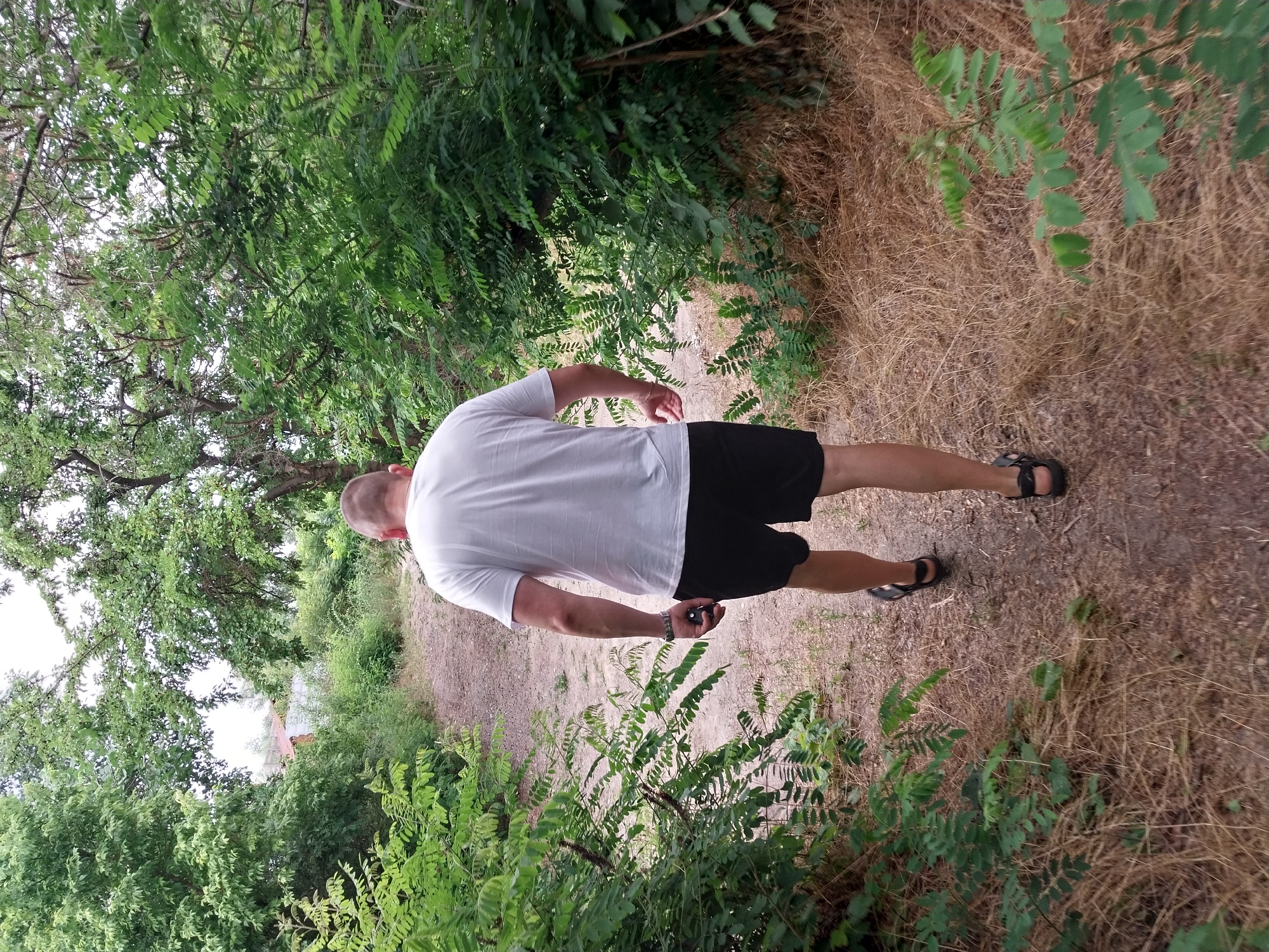 A man wearing a white t-shirt and black shorts walks away from the camera on a dirt path surrounded by lush green vegetation. The path appears to be in a natural or semi-wild area, possibly a park or a rural setting. The trees are dense, with many of them overhanging the path. The ground is covered with dry grass and leaves in some areas. The sky is not visible in the frame. The scene is static, with no visible interactions between the man and any other people or animals. The lighting suggests it is daytime. This scene could be located in the city of Stari Kodaky, Ukraine, given its naturalistic setting.