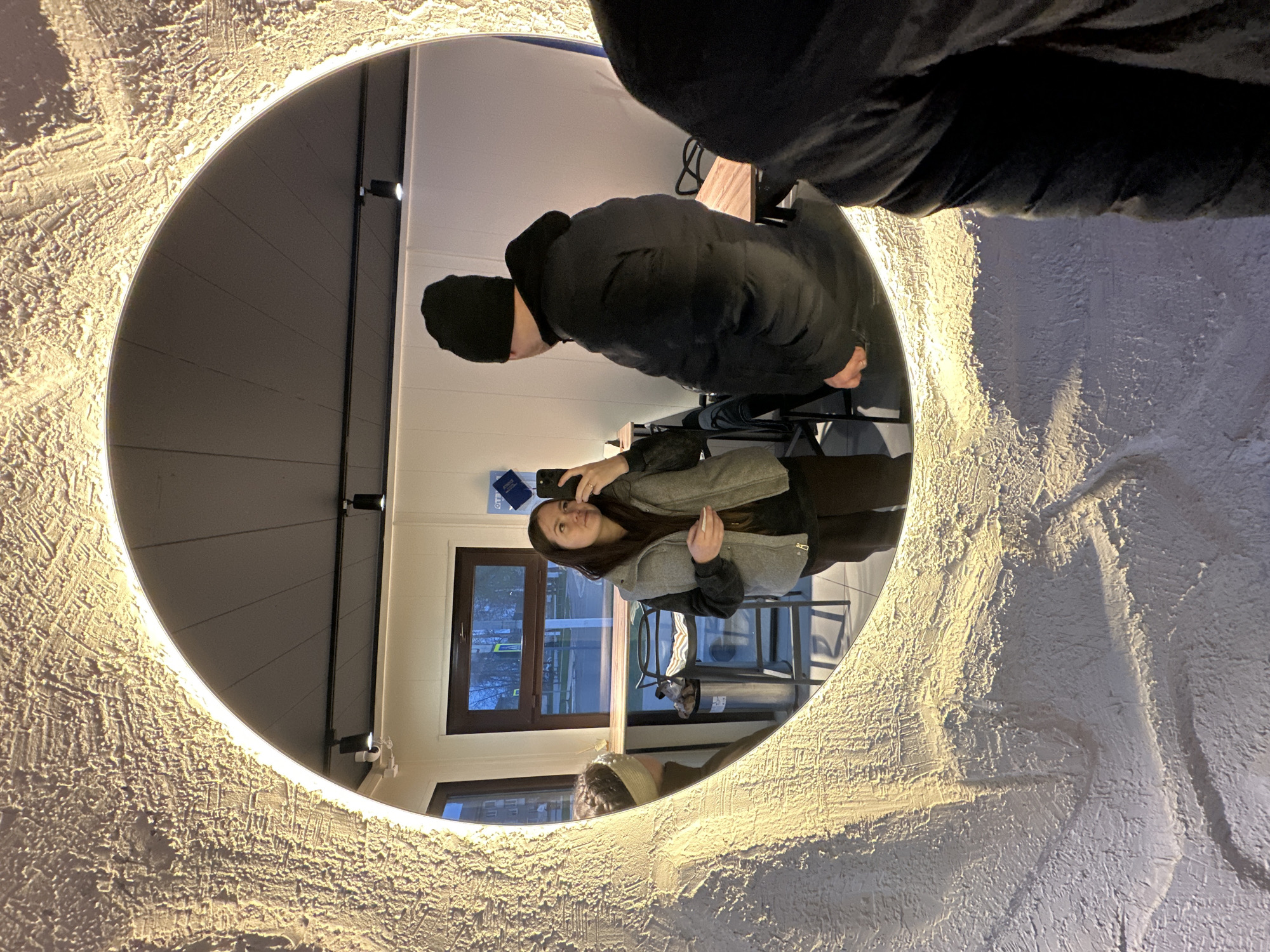 A mirror reflects two people and their surrounding environment. The mirror is circular with a textured white frame and a light around its inner edge. A person wearing a black beanie and a black jacket is reflected at the top of the mirror. A woman wearing a gray jacket is reflected below him, appearing to hold a cell phone. Outside the mirror is a building with windows. The reflection shows the building's interior, including chairs and a garbage can. The location is Chaikovsky, Russia.