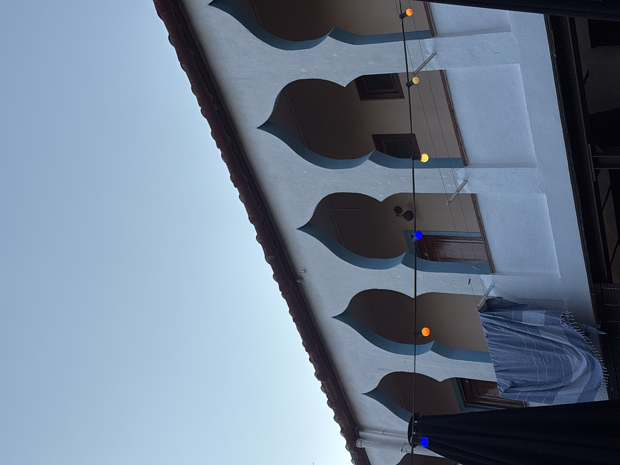 This image depicts the exterior of a building with distinctive arched windows and a tiled roof, characteristic of Mediterranean architecture. The building is painted white with blue trim around the arches. A string of colored lights, including blue and orange bulbs, is visible. A blue and white striped towel is hanging from a line on a balcony. A large black fabric object, possibly an umbrella or awning, is visible on the left side of the frame. The sky is clear and blue. The architectural style and the presence of a tiled roof suggest a location in Palafrugell, Spain, a town known for its whitewashed buildings and coastal influences.