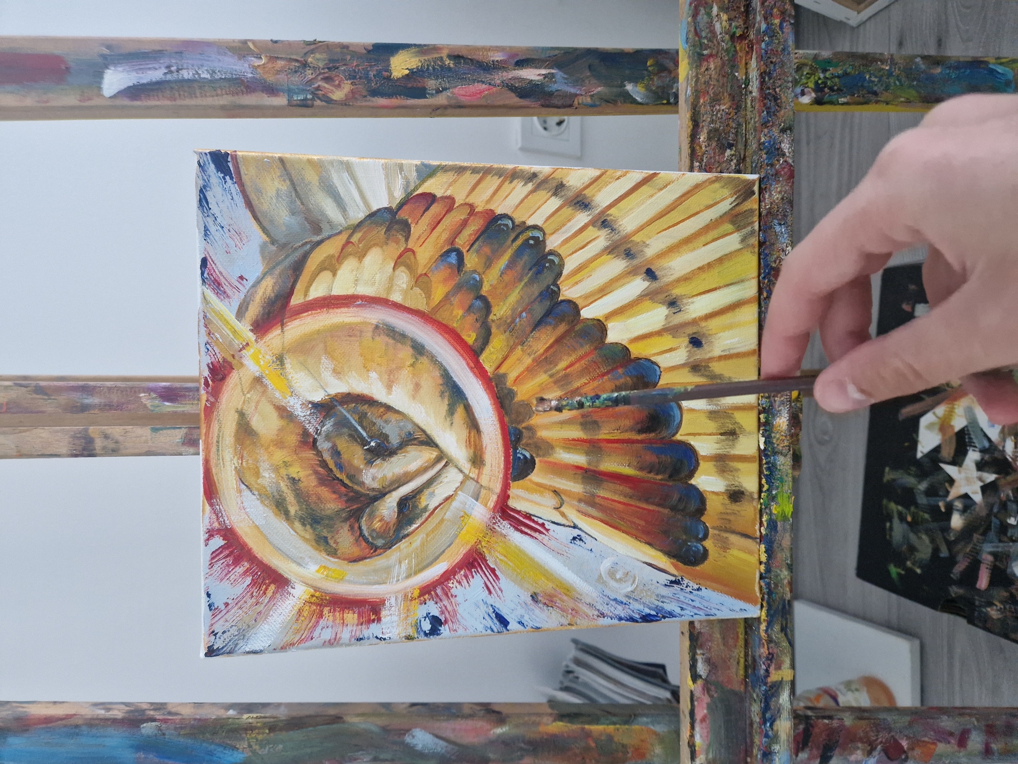 An overhead, slightly angled shot captures a person actively painting on a canvas. The artwork is oriented horizontally on a paint-splattered wooden easel, showing a vibrant, abstract or semi-abstract design. The painting is dominated by warm colors such as yellow, orange, and red, accented with blues and whites. A central circular motif, resembling an eye or a stylized organic form, is encircled by radiating, feather-like or petal-like shapes. These radiating elements transition from lighter yellows at their base to darker oranges, reds, and blues at their tips, displaying prominent brushstrokes and texture.
A light-skinned hand, likely the artist's, holds a thin paintbrush with a green-tipped brush, positioned near the right side of the canvas as if adding fine details to the radiating forms. The wooden easel supporting the canvas is visibly stained with a multitude of dried paint colors, indicating frequent use. In the background, a plain white wall features an electrical outlet, suggesting an indoor setting, possibly an art studio or a dedicated creative space. Parts of other canvases, a wooden floor, and a stack of papers or magazines are also visible, reinforcing the artistic environment. There is no visible text in the image.