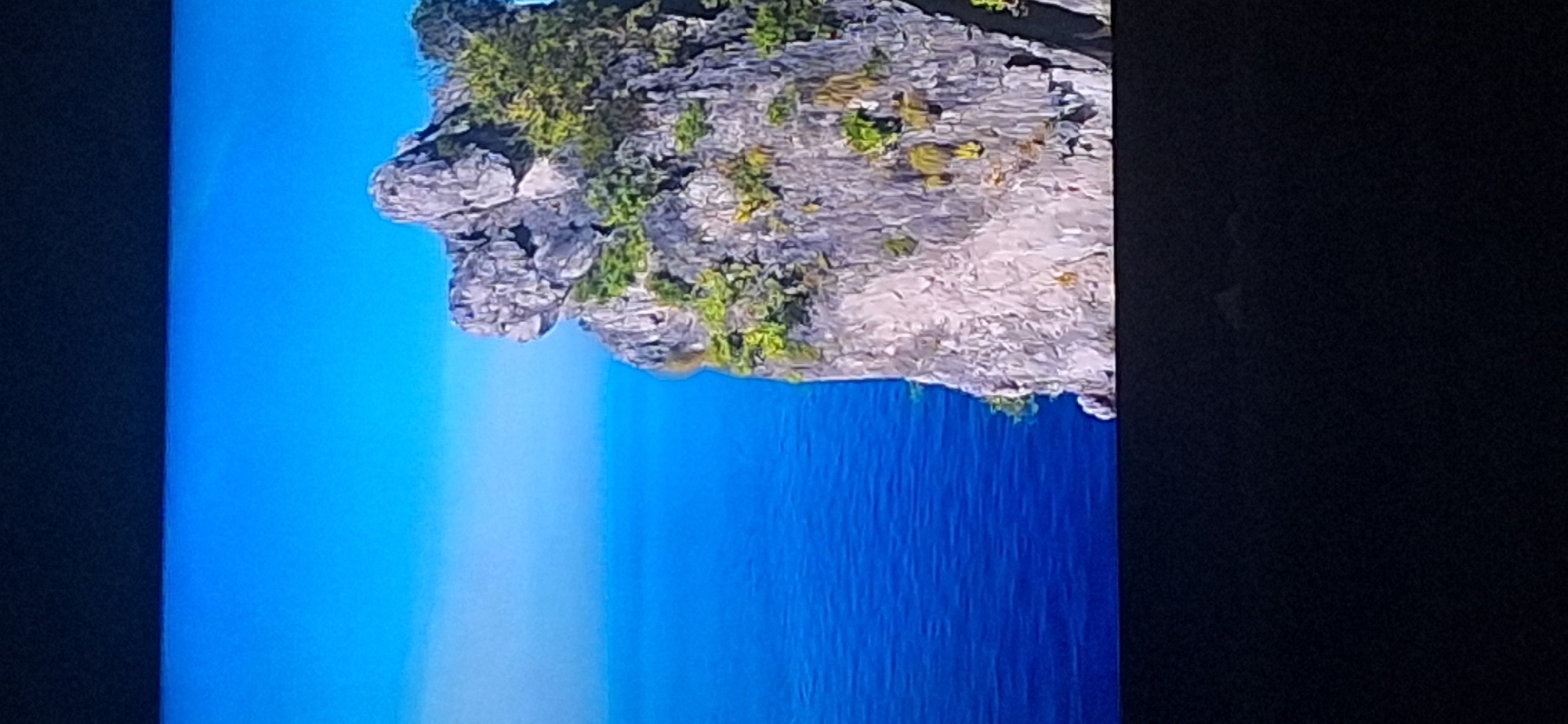 The image shows a rocky cliff face rising from the ocean under a bright blue sky. The cliff is covered in patches of green vegetation. The water is a deep blue. The bottom of the image is black, suggesting it is a photograph of a screen. There is no indication of the city of Bila Tserkva, Ukraine.