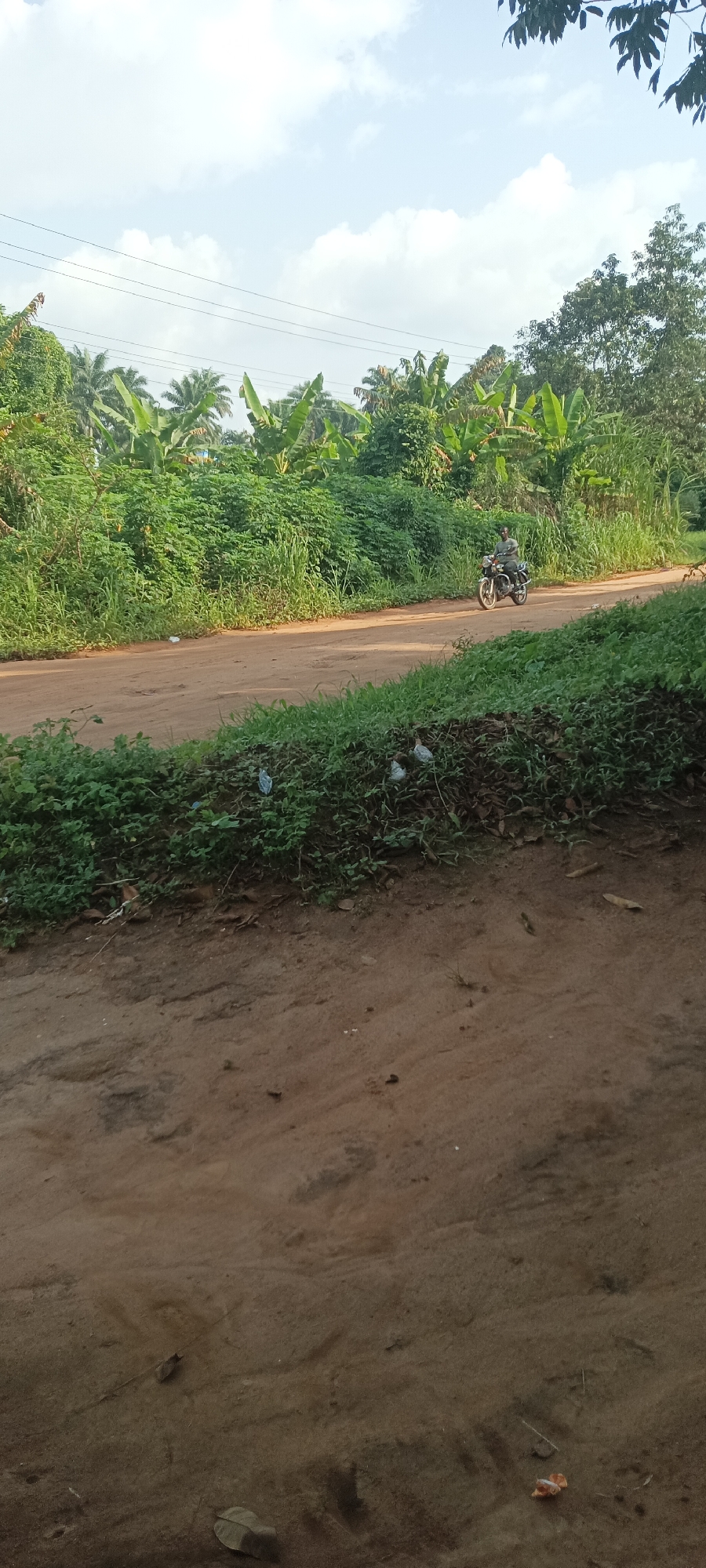 The image depicts a dirt road in Amauzu, Nigeria, lined with dense vegetation. A man rides a motorcycle on the road. The foreground shows a dirt patch with scattered debris. Overhead, power lines are visible against a partly cloudy sky.