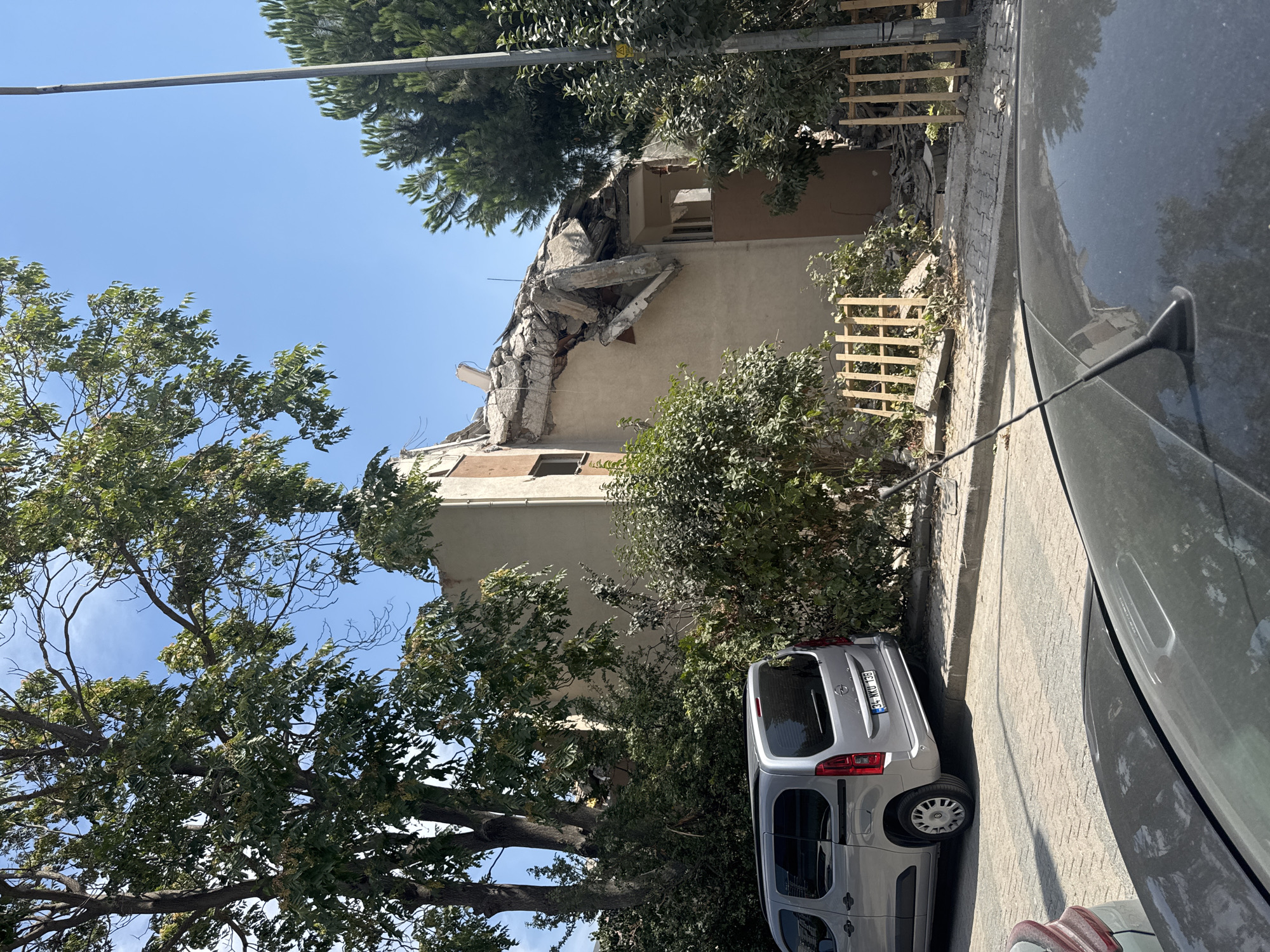 The image shows a street scene in Gungoren, Turkey, with a severely damaged building. The building's roof has collapsed, and debris is scattered around its base, indicating structural failure. Several trees and bushes are growing around the building and on the sidewalk. A silver van is parked on the street in front of the damaged structure, and a portion of another car is visible in the foreground. The sky is clear and blue, suggesting fair weather. The scene appears to be in a residential or mixed-use area of the city.