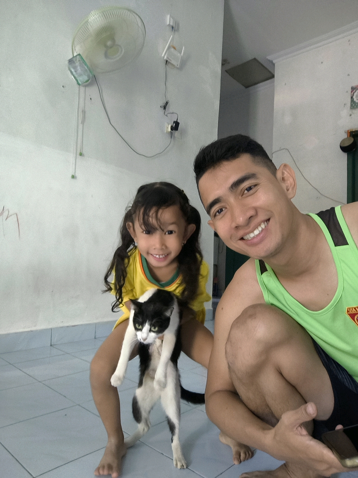 In an indoor setting, a man and a young girl are posing for a selfie with a black and white cat. The man, wearing a green tank top, is crouched down and smiling at the camera. The girl, dressed in a yellow shirt, is holding the cat, which is suspended vertically. The background features a white wall with a ceiling fan and electrical outlets. The flooring appears to be tiled. The overall impression is a casual, domestic scene, possibly in a home in an urban area of Indonesia.