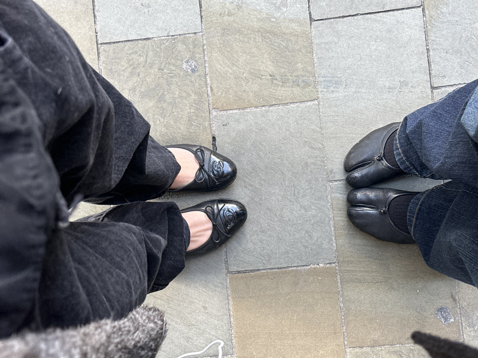 An overhead photograph depicts two pairs of feet standing on an outdoor paved surface in London, United Kingdom. In the upper portion of the frame, a person is shown wearing dark grey, possibly denim, wide-leg trousers and black leather Chanel ballet flats. These flats feature a bow and an embossed interlocking 'C' logo on the patent leather toe cap. In the lower portion of the frame, a second person wears dark blue denim jeans, black socks, and black tabi-style shoes, characterized by a split-toe design and a small circular button closure. The ground is composed of irregularly shaped rectangular paving stones in shades of light grey and tan. A thin white cord is partially visible on the far left, adjacent to the upper person's leg. An elongated shadow is cast on the lower left of the pavement. The precise location within London is not identifiable from the visual evidence presented.