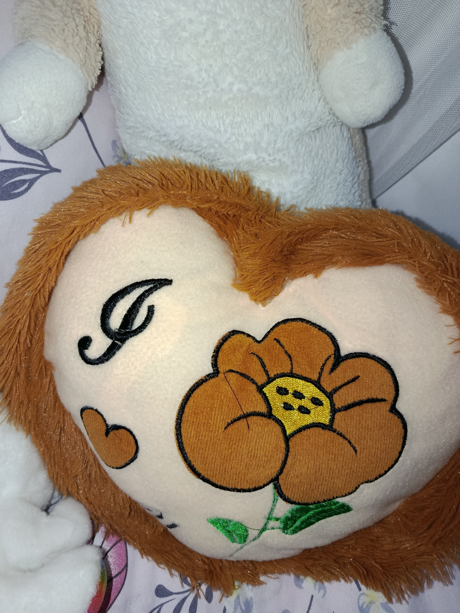 The image is a close-up, top-down view of a plush heart-shaped pillow. The pillow is light beige with a fuzzy brown border. Embroidered on the pillow is a large, stylized brown and yellow flower with green leaves. To the left of the flower, there is a black embroidered cursive letter "I" and a small heart shape. The pillow is lying on a surface with a light-colored fabric featuring a subtle floral or leaf pattern in a darker shade. In the background, parts of two other plush toys are visible: one is white and fuzzy with beige accents, and another appears to be a lighter color. The lighting is dim, creating some shine on the fuzzy textures. There is no indication of the location being in Tashkent, Uzbekistan, or any specific city.