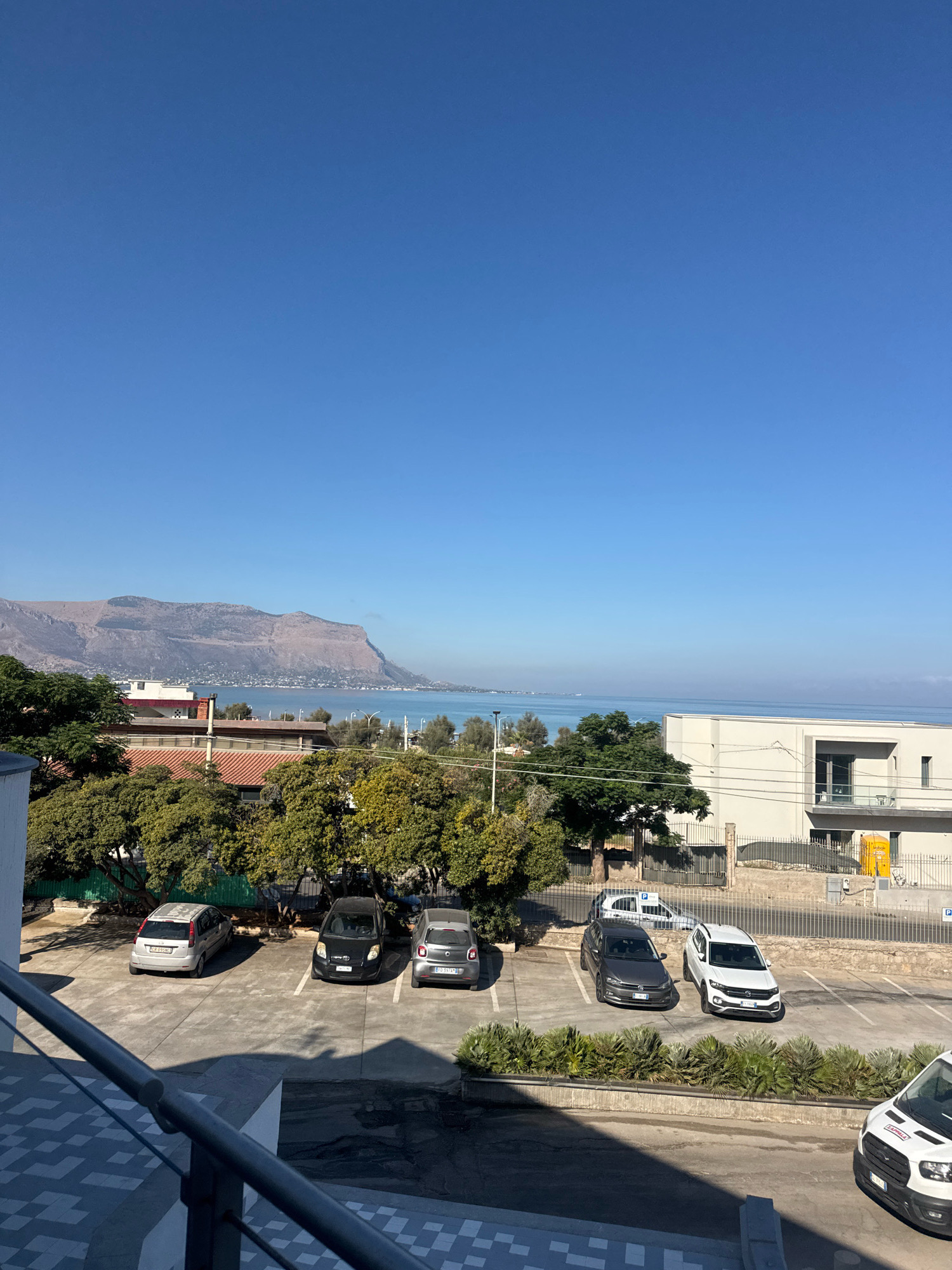 A clear, bright sky dominates the upper portion of the image. Below the sky, a hazy blue body of water, likely the Tyrrhenian Sea, stretches across the horizon. In the distance, a large, rugged mountain range rises from the coastline. The middle ground features a coastal town with low-rise buildings, interspersed with lush green trees. A distinct feature of the landscape is a road with streetlights running along the waterfront. In the foreground, a parking lot is visible with several cars parked. The parking lot is bordered by a modern building with white walls and dark-framed windows. A yellow portable toilet is situated near the building. The image appears to be taken from an elevated position, possibly a balcony, overlooking the parking lot and the scenic view beyond. The presence of the characteristic mountain range and coastal setting strongly suggests the location is within or very near the town of Capaci, Italy, a region known for its beautiful coastline and proximity to prominent geological formations.