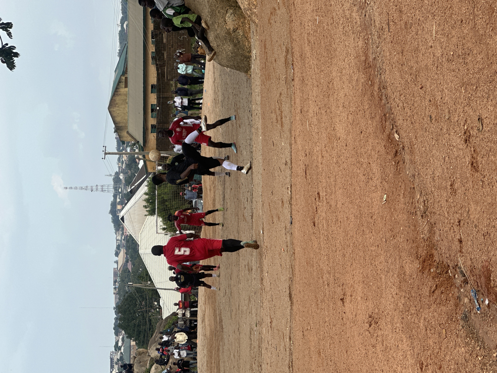 The image shows an outdoor football match in progress on a dirt field. Several players are visible on the field, some in red jerseys and others in black. One player in a red jersey with the number "5" prominently displayed on his back is a central figure, appearing to be engaged in the game. Spectators are gathered along the sidelines, some sitting on rocks. In the background, buildings and infrastructure are visible, suggesting an urban or semi-urban setting. The sky appears overcast. The scene is likely taking place in Jos, Nigeria, as indicated by the location context. There is no visible text in the image. The overall atmosphere seems to be one of energetic sporting activity and community engagement.