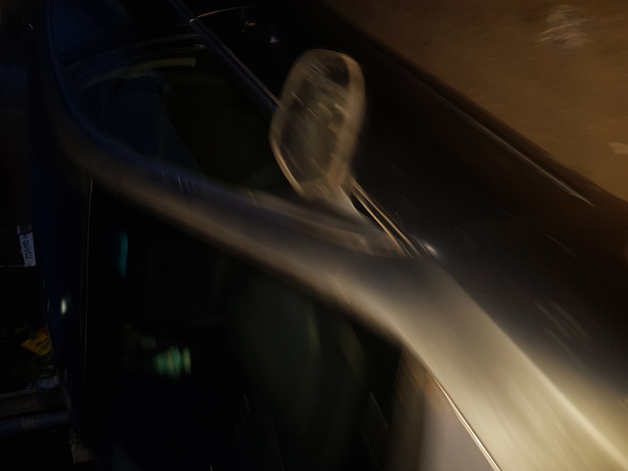 The image shows a blurred and rotated view of a car. The scene is dimly lit, possibly at night. The car's side mirror is visible, but blurry. The car's paint appears dark, possibly black or dark blue. The background is out of focus and appears to be an urban environment. The image shows items along the bottom left. The image is too blurry to determine exact location in Shangisha, Nigeria.
