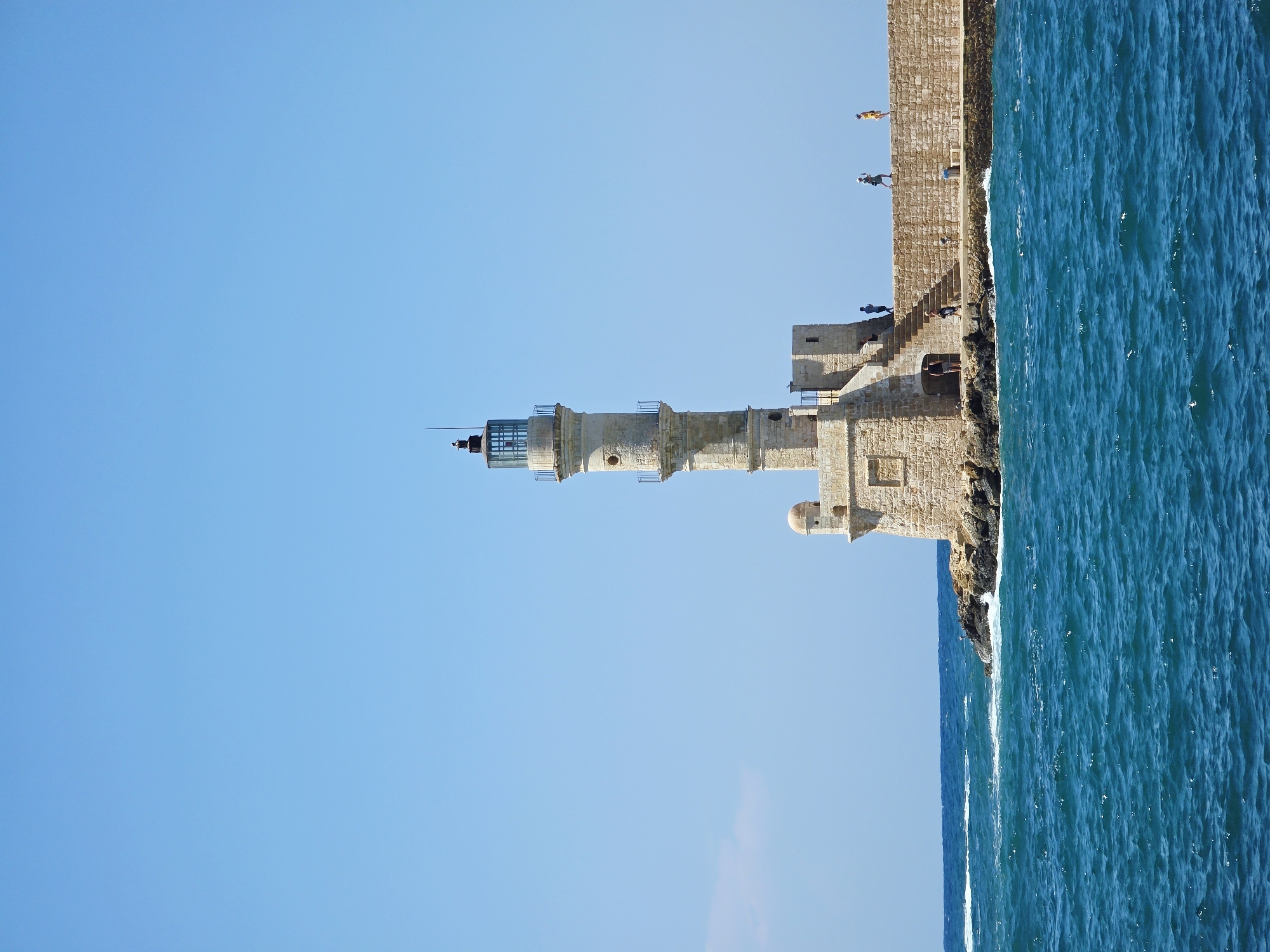 The image shows the Venetian lighthouse in Chania, Greece, standing tall against a clear blue sky. The lighthouse, constructed of stone, has a cylindrical tower with multiple levels, topped by a lantern room. A stone walkway, also made of blocks, extends from the lighthouse into the bright blue sea, which is choppy with white-capped waves. Several people are visible on the walkway, some walking, others standing. The lighthouse is situated on a rocky outcrop. The atmosphere is bright and sunny, suggesting a warm day.