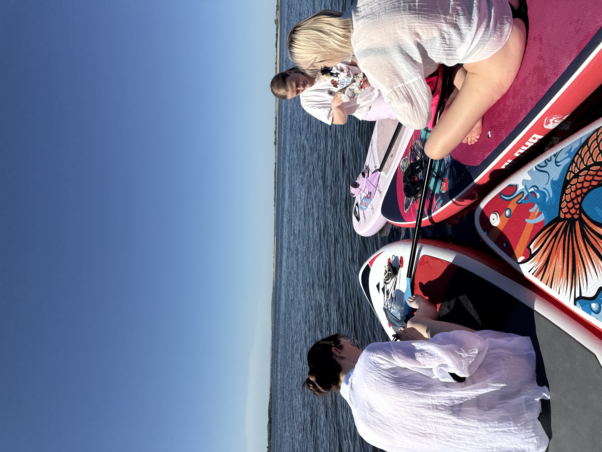 The image captures four women on paddleboards in what appears to be a body of water. The sky is clear and blue, suggesting a sunny day. The water stretches towards the horizon with a thin strip of land visible in the distance.
The women are dressed in light-colored clothing, with at least three wearing white tops. They are positioned on several paddleboards. One board has a design featuring what looks like an anime character and other designs, and another has a design with scales and what resembles the tail of a fish. A woman in the foreground is wearing sunglasses and has her hair up. The women appear to be interacting and looking at something, possibly food. The scene suggests a recreational outing on the water. The location is likely Усть-Курдюм, Russia.