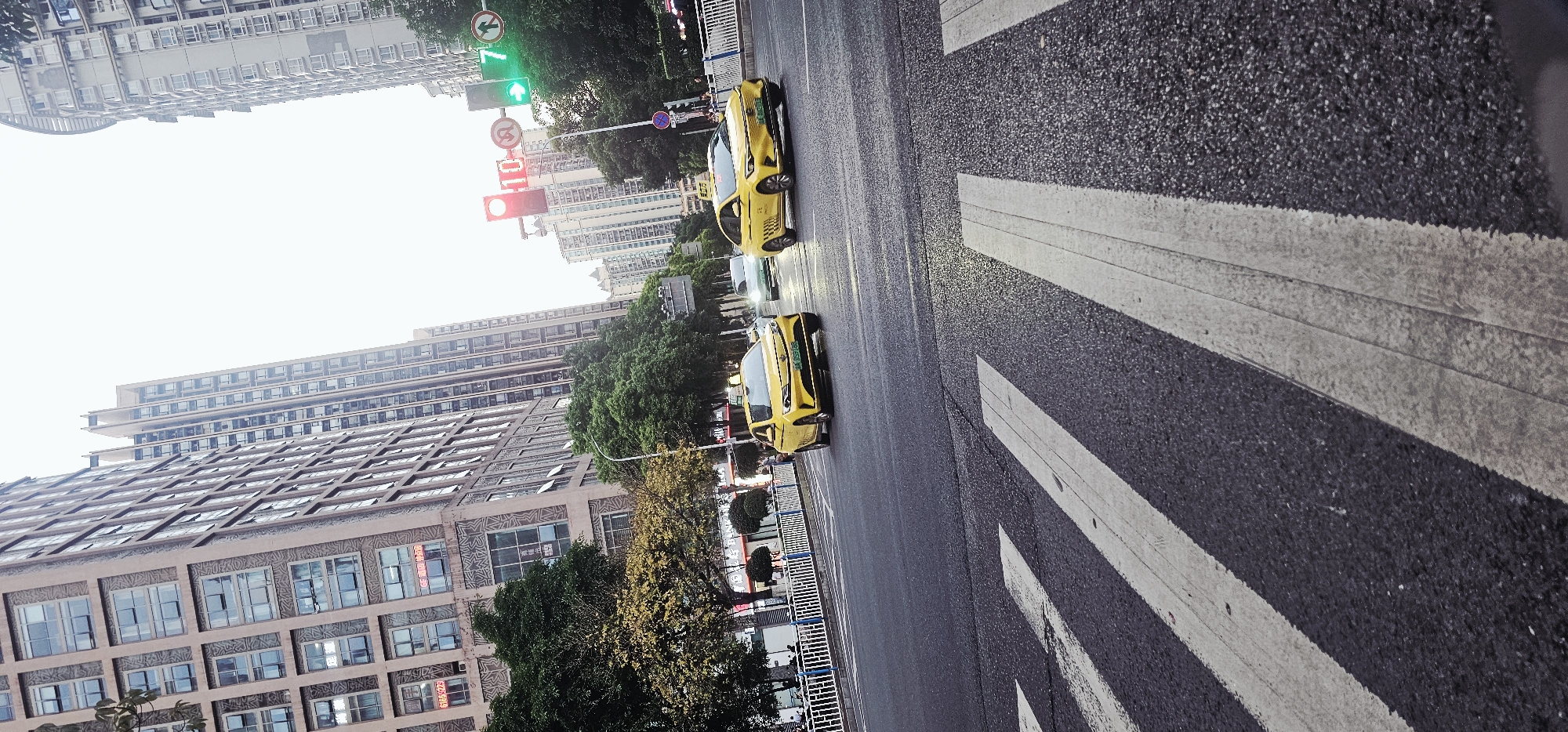 The image shows a street scene in Yuzhong District, China. Two yellow taxis are driving on the road. There is a crosswalk with white stripes in the foreground. Buildings are visible on both sides of the street, with trees lining the road. A traffic light displays a green light in one direction and a red light in the other. The perspective is from a low angle, and the image is rotated slightly.