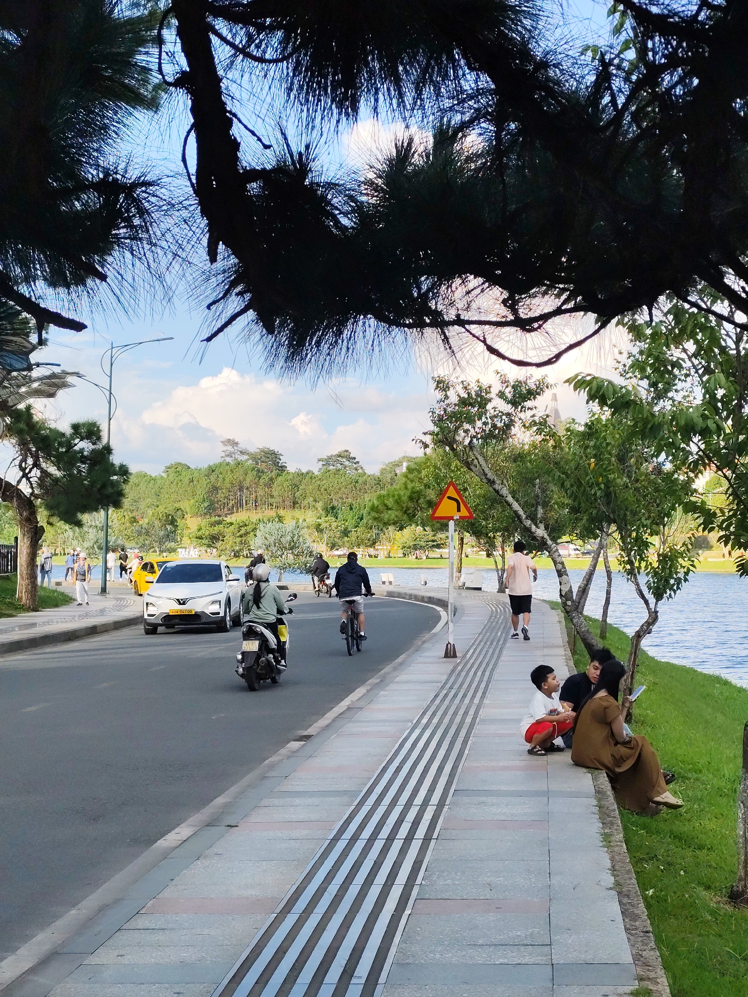 The image depicts a scenic view of Xuân Hương Lake in Đà Lạt, Vietnam. On the left side of the frame, a paved road runs alongside a sidewalk. A white car is parked on the road, and several people are walking on the sidewalk. A motorcycle is also visible on the road. The sidewalk is bordered by a grassy area that slopes down to the lake. In the background, lush green trees and hills surround the lake, and the sky is partly cloudy. On the right side of the frame, a group of people are sitting on the grass by the lake, some of whom appear to be looking at a phone.