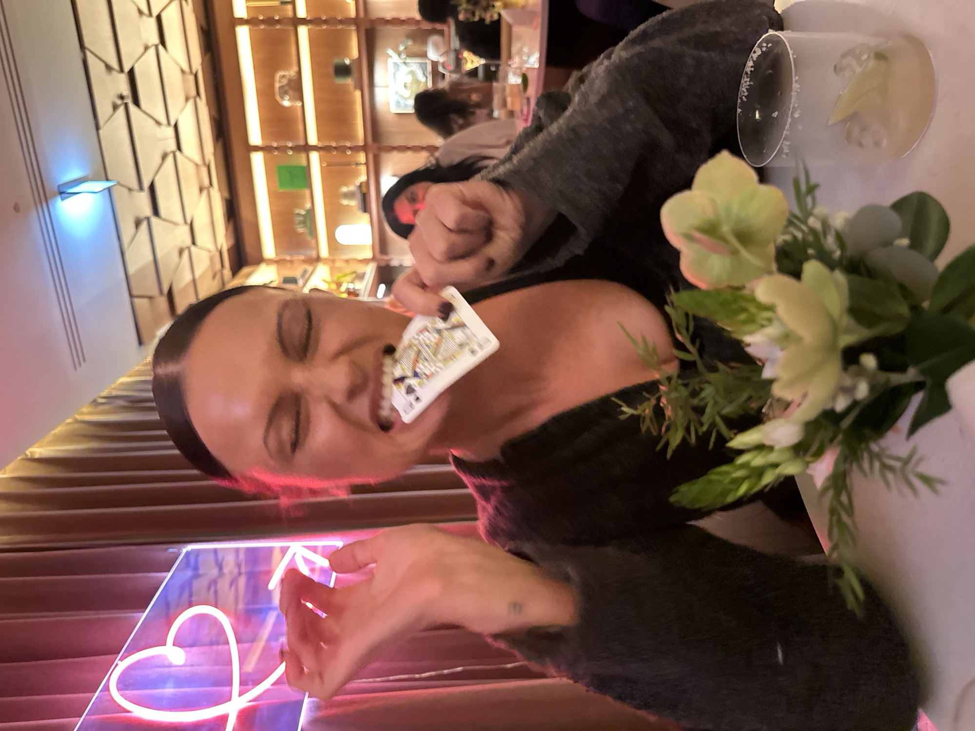 This vertically oriented image captures a playful moment with a woman posing in an upscale indoor setting, likely a restaurant or bar in London, UK. The primary subject is a woman, shown from the chest up, with dark hair pulled back. She makes a humorous, exaggerated expression, holding a playing card—the Queen of Clubs—partially in her teeth, with her eyes squinted shut. She is wearing a dark, possibly knit, top. Her left hand is visible near a glowing pink neon sign, which features a heart shape. A small, subtle tattoo can be seen on her left wrist.
To her right, on a table, a cocktail in a stemmed glass with a sugared rim is partially visible, positioned behind an artificial floral arrangement of light green and white flowers with lush greenery. The background features warm-toned wooden shelving filled with various decorative items, and walls with a distinct geometric, textured pattern. Blurred figures in the background suggest other patrons. The lighting is artificial and warm, indicating an evening setting. The overall atmosphere appears social and lighthearted.