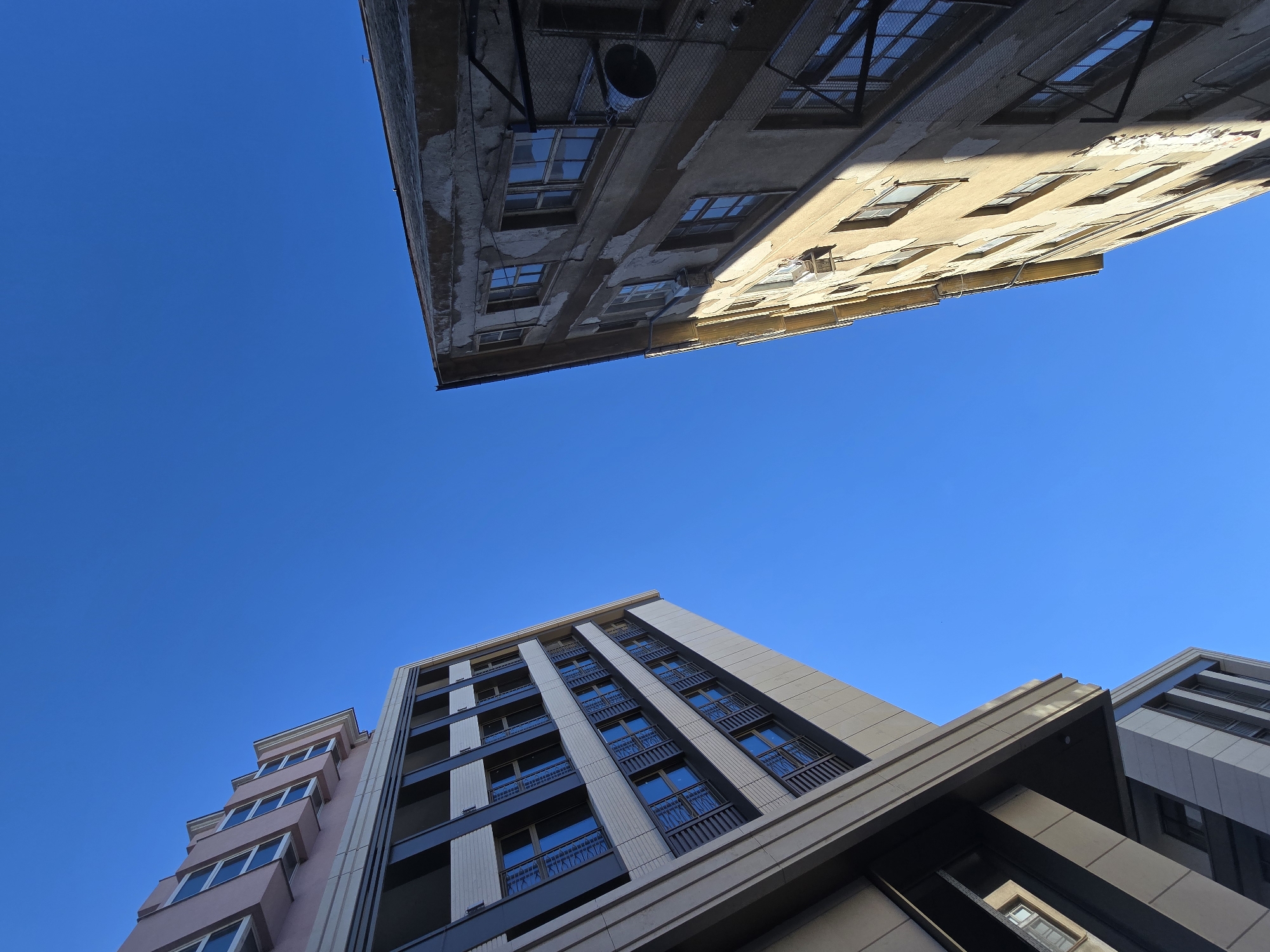 The image presents a low-angle view of several buildings against a clear blue sky. The building at the top of the frame is older, with a weathered facade, visible cracks, and a mix of windows. It has an indistinct grey color. The buildings at the bottom of the frame are more modern, with a mix of pink, white, black, and beige, and a greater use of contrasting materials. The photo is taken outdoors, with a very blue cloudless sky. The exact city and location are not discernible from the image.