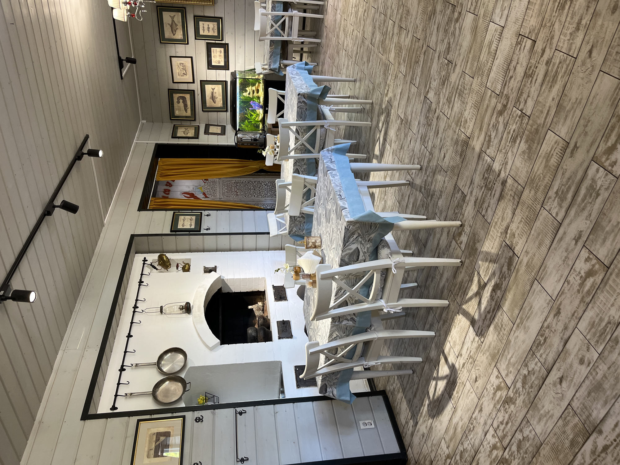 The image depicts the interior of a restaurant located in Ново-Никольское, Russia. The dining area features several tables covered with patterned tablecloths, surrounded by white wooden chairs with cross-back detailing. One table in the foreground is set with a salt and pepper shaker, a napkin holder, and a small vase with a white flower.
The walls are constructed of light-colored wooden planks. A large, white-tiled fireplace with an arched opening dominates one section of the wall. Above the fireplace, a black metal rack holds two large pans and a lantern. Various framed pictures, some depicting fish, hang on the walls.
To the right of the fireplace, a large aquarium containing fish and aquatic plants is visible. Several tables and chairs are arranged around the aquarium, suggesting it's a focal point of the dining space. The floor is made of light-colored wooden planks, with shadows cast by the furniture. The overall lighting appears to be a combination of overhead track lighting and possibly natural light from unseen windows. No people are present in the image.