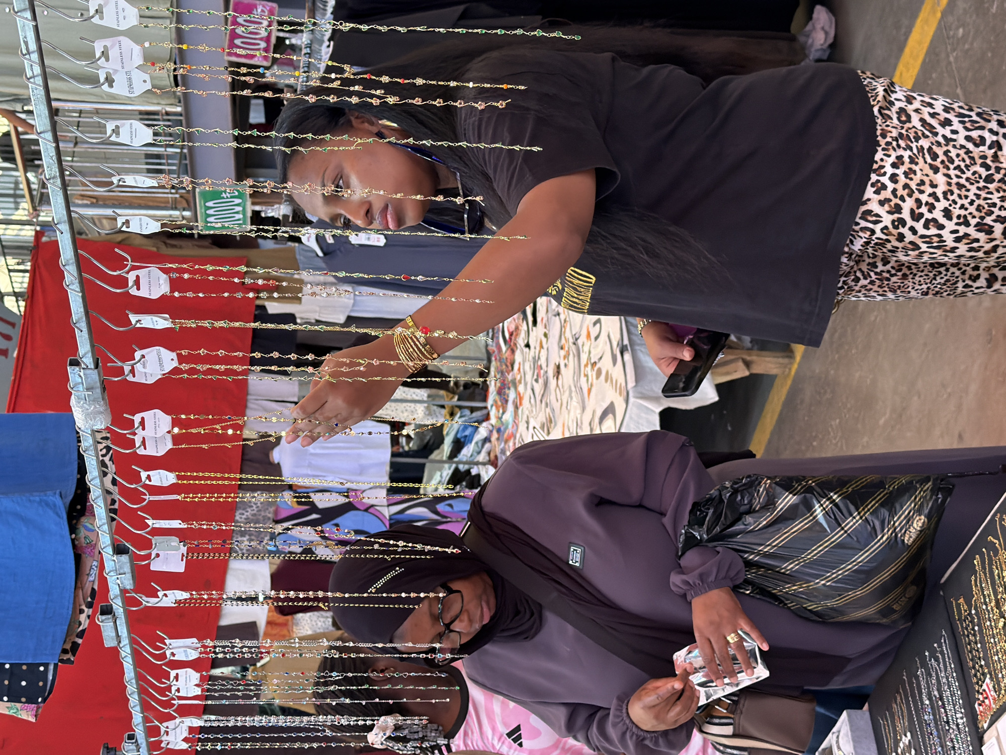 Two women are seen at a market stall selling beaded necklaces. The woman on the left, wearing a dark purple hijab and glasses, is examining a clear plastic package of jewelry. She is wearing a dark purple long-sleeved shirt and carrying a black and gold striped plastic bag in her right arm. In front of her, on a black display mat, are rows of necklaces and bracelets.
The woman on the right is wearing a black t-shirt and leopard-print pants. She has long, dark hair and is looking at the necklaces hanging from a metal rack above. She is reaching out with her right hand, possibly to select or examine a necklace. She is wearing several gold bracelets on her left wrist.
Behind the women, a red fabric is visible, possibly part of the stall's awning. Signs with Turkish currency symbols (₺) and numbers are hanging among the necklaces. The overall environment suggests an outdoor market setting, potentially in Kadıköy, Turkey, given the presence of Turkish currency and the commonality of such street markets in the city. The floor is concrete, with a yellow line marking a boundary. Other people and stalls are visible in the background, indicating a bustling market.