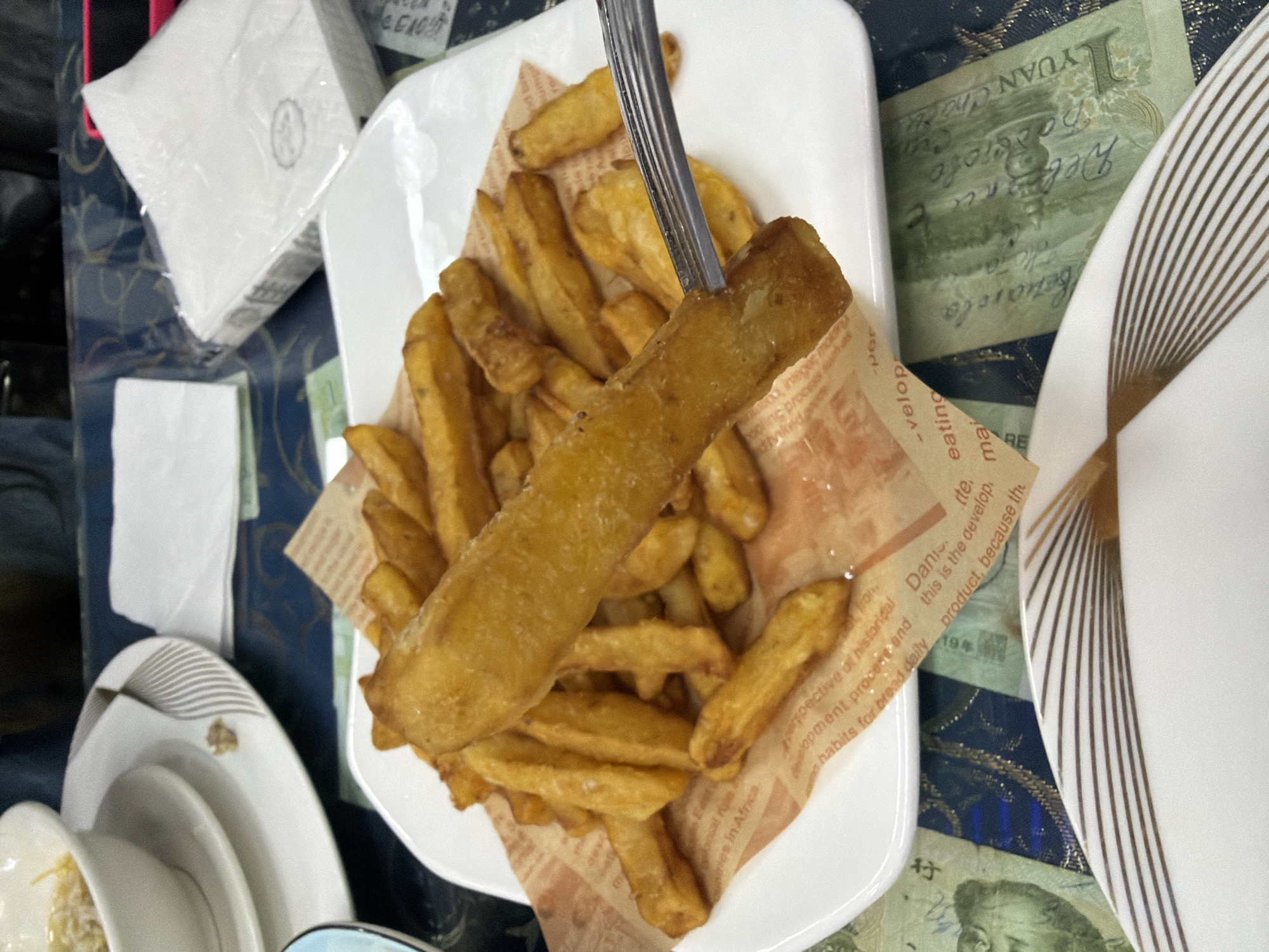 A top-down shot captures a plate of golden-brown french fries (chips) served in an indoor setting, likely a restaurant or cafe. A large piece of fried food, possibly fish, is impaled on a silver fork and held above the generous portion of fries. The fries are served on a white rectangular plate, lined with a piece of brown greaseproof paper featuring partially visible English text that seems to be part of an article or description, with phrases like "this is the develop" and "habits for bread daily" discernible.
The plate sits on a dark blue tablecloth with a subtle pattern. Scattered on the tablecloth are several banknotes. One visible green banknote clearly shows "1 YUAN" and Chinese characters, identifying it as Chinese Renminbi. Other banknotes are partially visible, one possibly depicting a portrait commonly found on Chinese currency.
Adjacent to the main dish, other white porcelain plates and bowls are visible, some stacked, and one with a decorative grey and gold linear pattern. A stack of individually plastic-wrapped white napkins, one bearing a small gray logo, is positioned in the upper left, with another unwrapped napkin below. The scene suggests a casual dining experience in Tongjiang, China, with the currency acting as a strong location cue. No people are visible, and the time of day is not indicated.