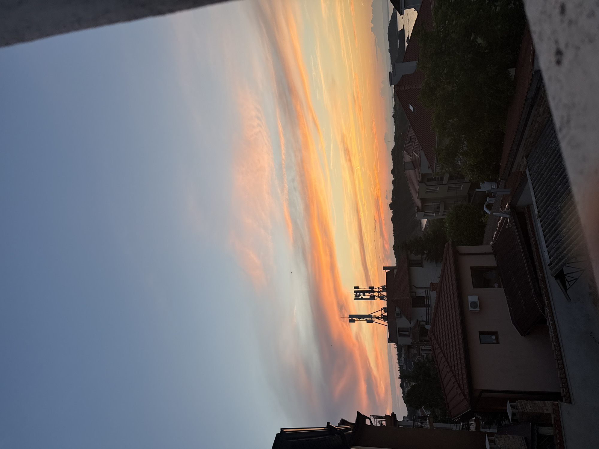 This is a photograph of a sunset over the city of Chernomorets, Bulgaria. The sky is filled with vibrant colors of orange, pink, and yellow, with wispy clouds stretching across the horizon. In the foreground, there are several buildings with red tile roofs, and on top of one building, there are two tall communication towers. The sea is visible in the distance, with a few rocky outcrops. The overall scene is peaceful and serene, with a sense of tranquility.